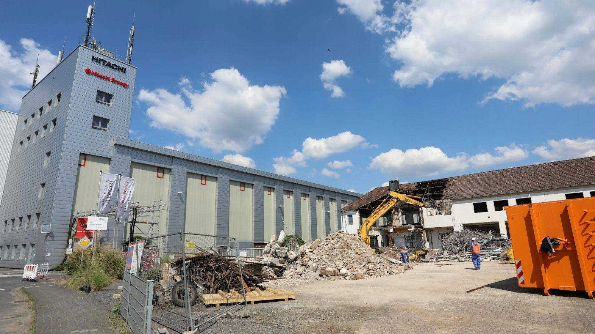 Neben einem Fabrikgebäude stehen ein Bagger und ein Container, am Rand liegt Bauschutt.