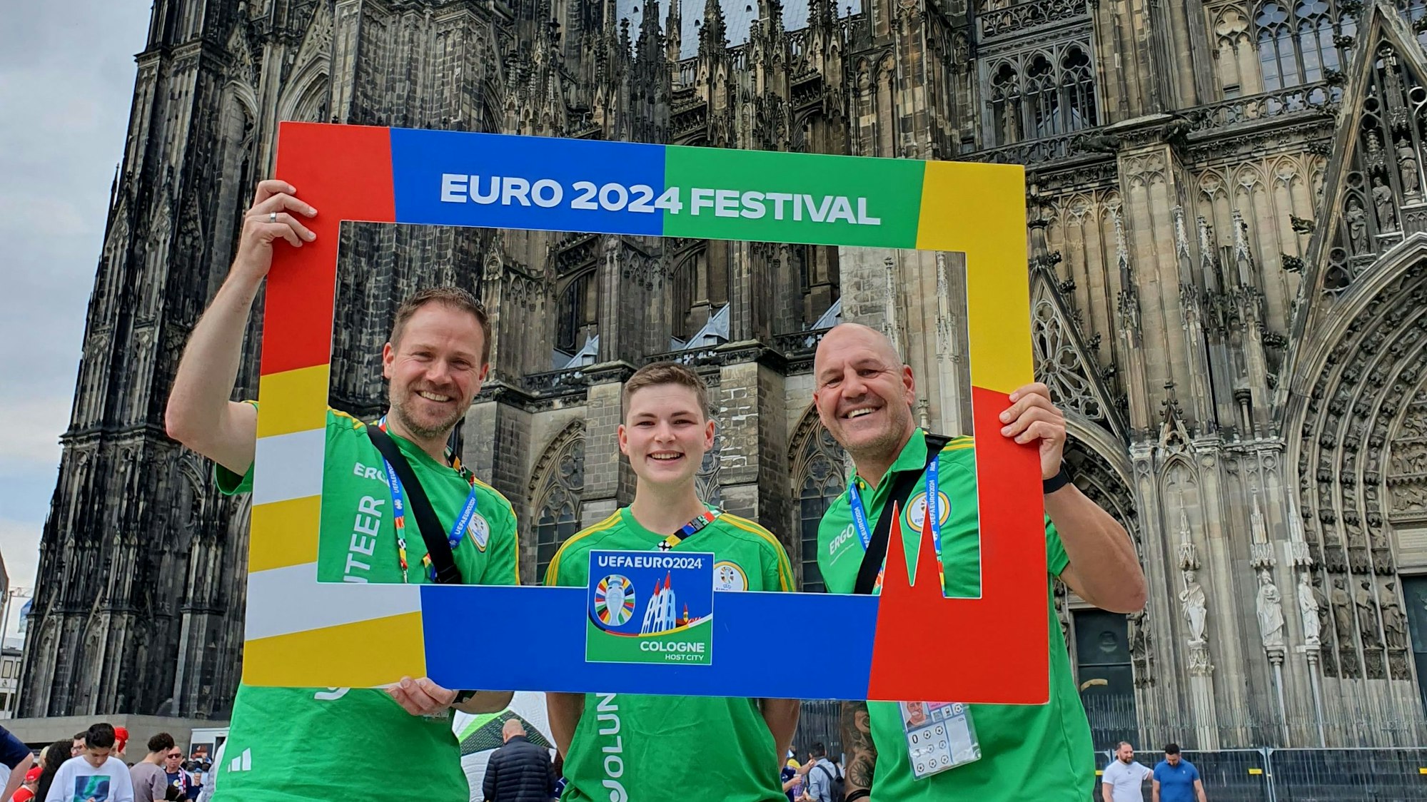 Die EM-Volunteers stehen vor dem Kölner Dom und halten einen Rahmen mit der Aufschrift „Euro 2024 Festival“.
