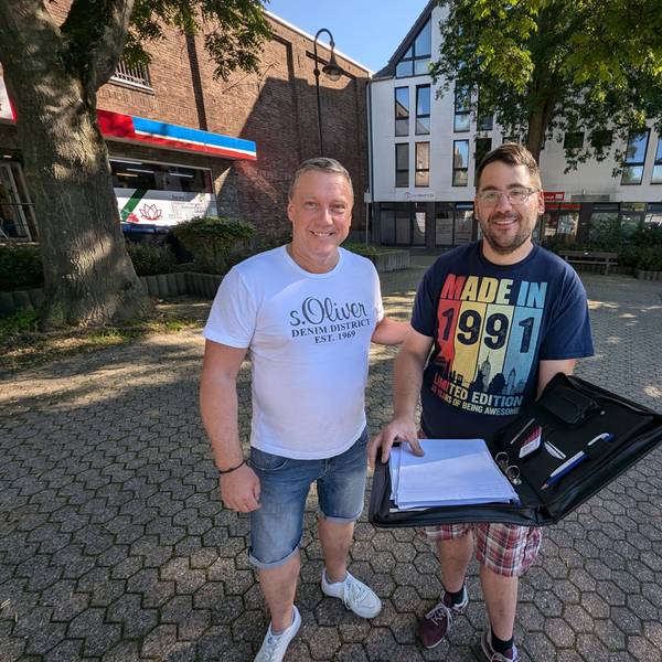 Zwei Männer stehen mit einer aufgeklappten Aktenmappe auf dem Platz an der Alten Synagoge in Alt-Hürth.