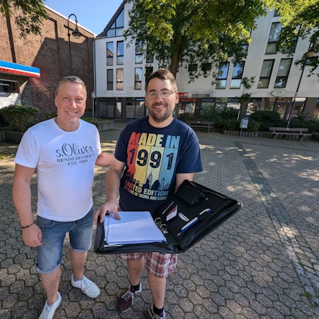 Zwei Männer stehen mit einer aufgeklappten Aktenmappe auf dem Platz an der Alten Synagoge in Alt-Hürth.