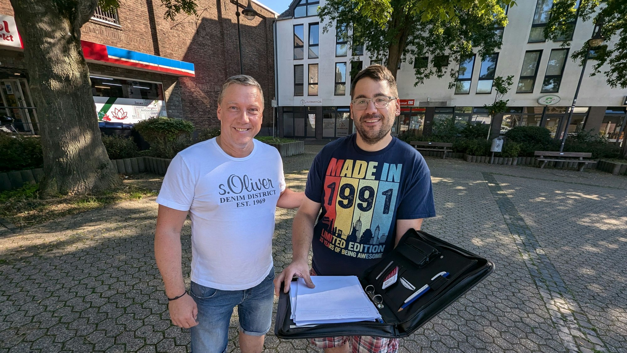 Zwei Männer stehen mit einer aufgeklappten Aktenmappe auf dem Platz an der Alten Synagoge in Alt-Hürth.