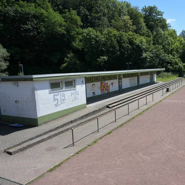 Rasenfläche und Aschenbahn eines Sportplatzes, daneben ein flacher Bau, in dem sich die Umkleiden befinden. Die Wände sind bemalt.
