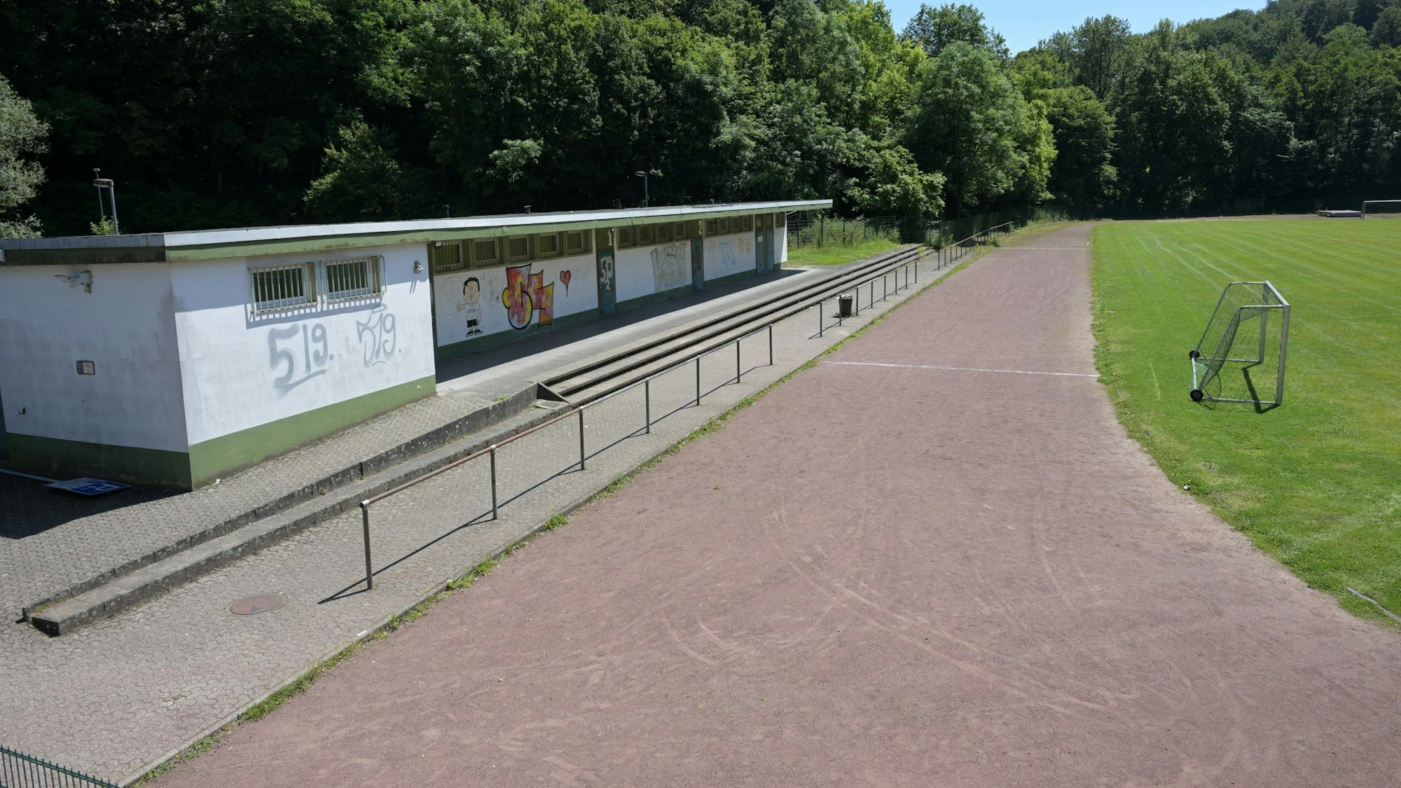 Rasenfläche und Aschenbahn eines Sportplatzes, daneben ein flacher Bau, in dem sich die Umkleiden befinden. Die Wände sind bemalt.