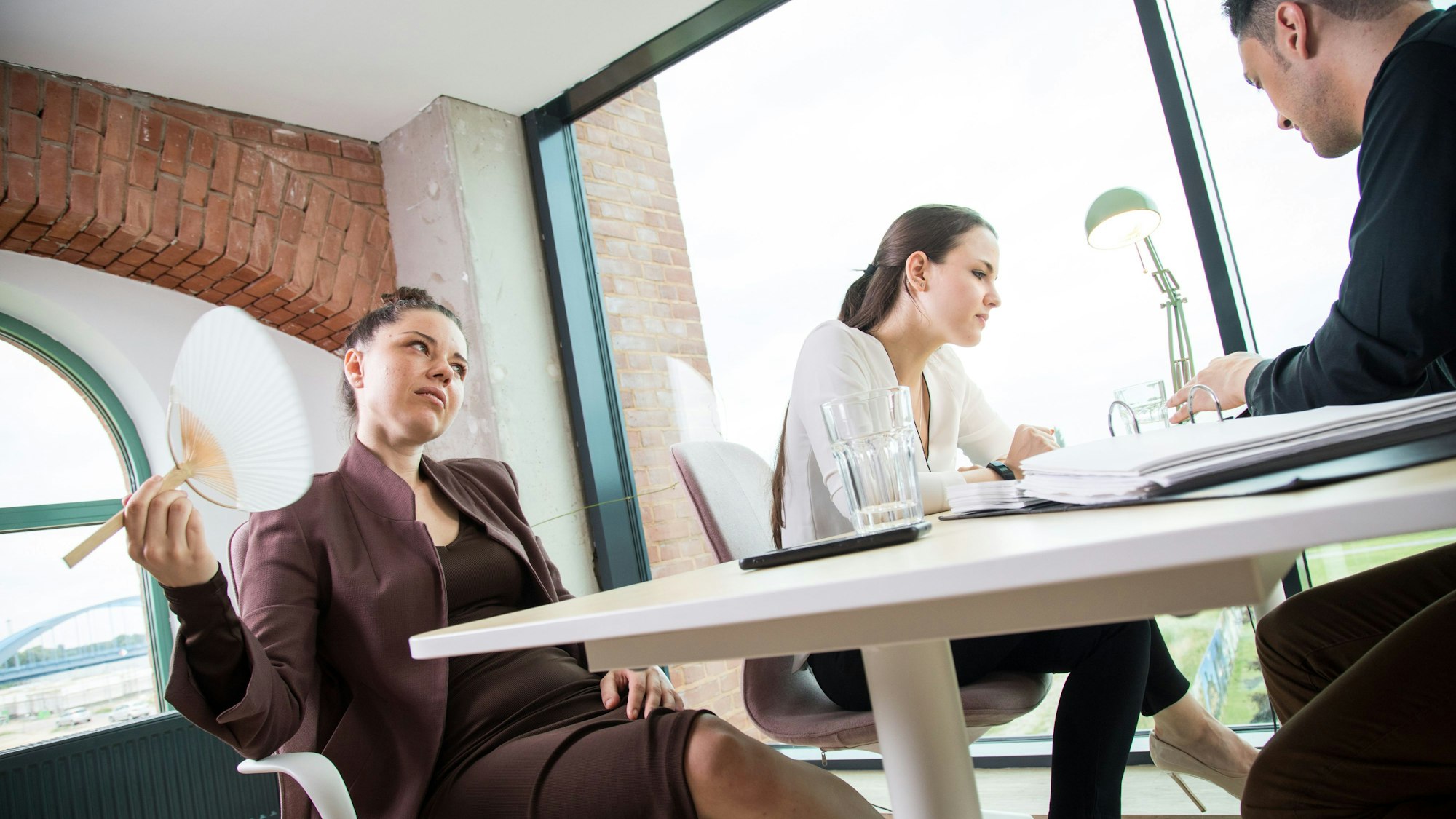 Drei Personen sitzen an einem Tisch. Eine Frau wedelt sich mit einem Fächer frische Luft zu.