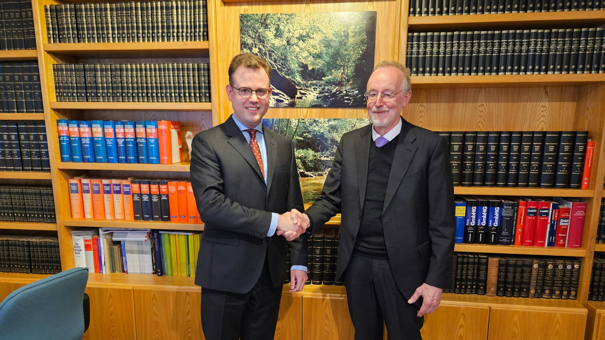 Wilhelm Scheuvens (r.) und Martin Strahl tragen dunkle Anzüge. Sie stehen vor einer Bücherwand und geben sich die Hand.