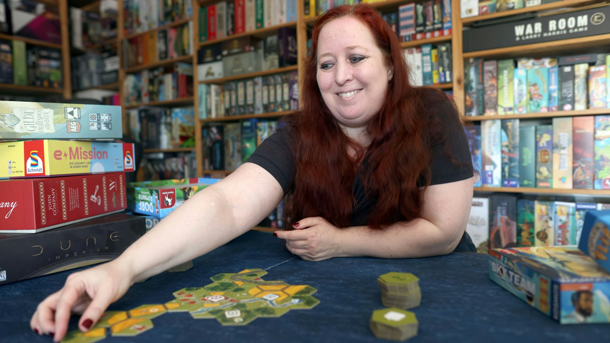 Martina Fuchs sitzt an einem Tisch und legt Spielkarten auf den Tisch. Hinter ihr im Regal und neben ihr auf dem Tisch stapeln sich Spiele.
