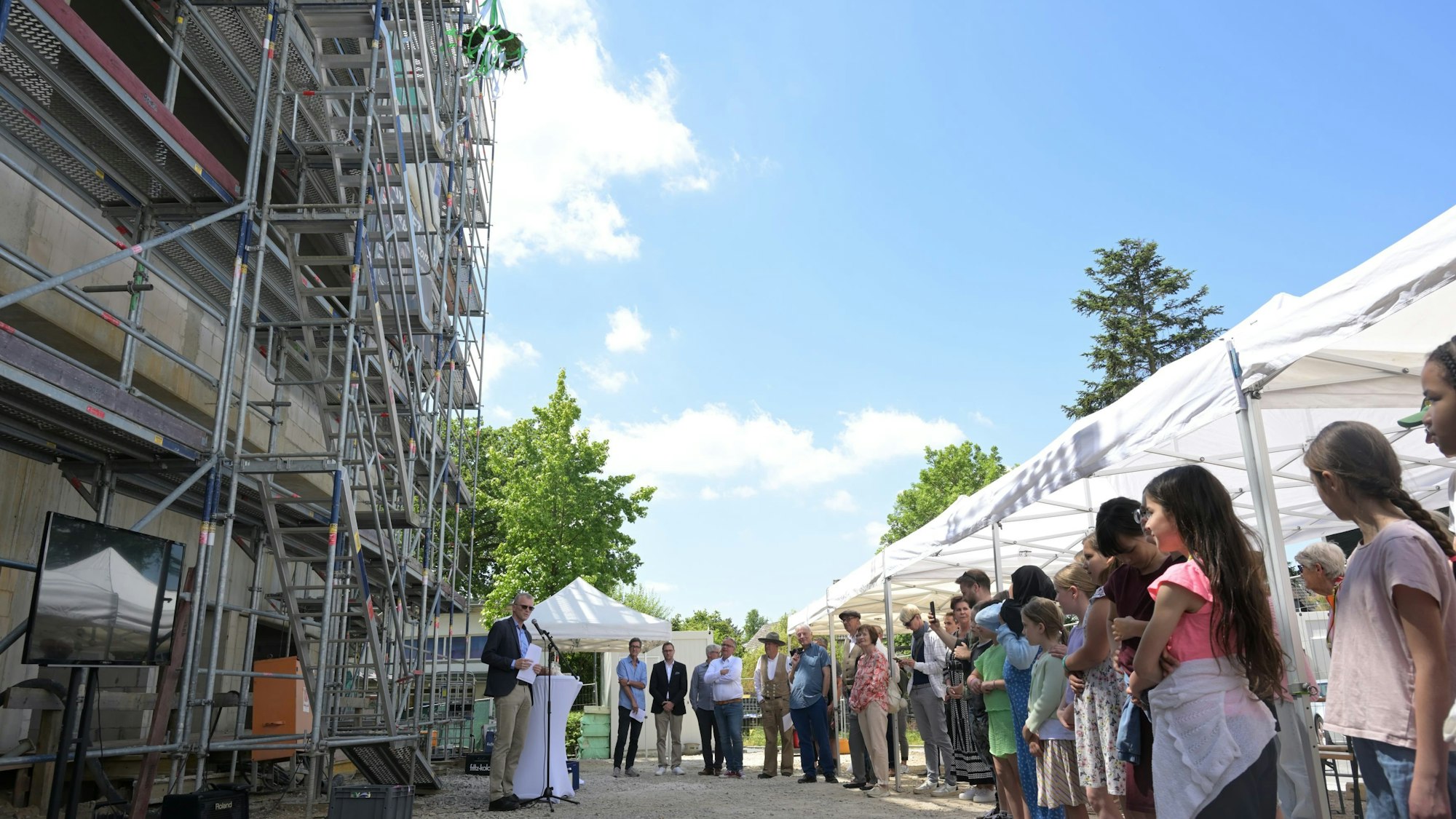Vor dem eingerüsteten Rohbau stehen Kinder, der Bürgermeister hält eine Rede.