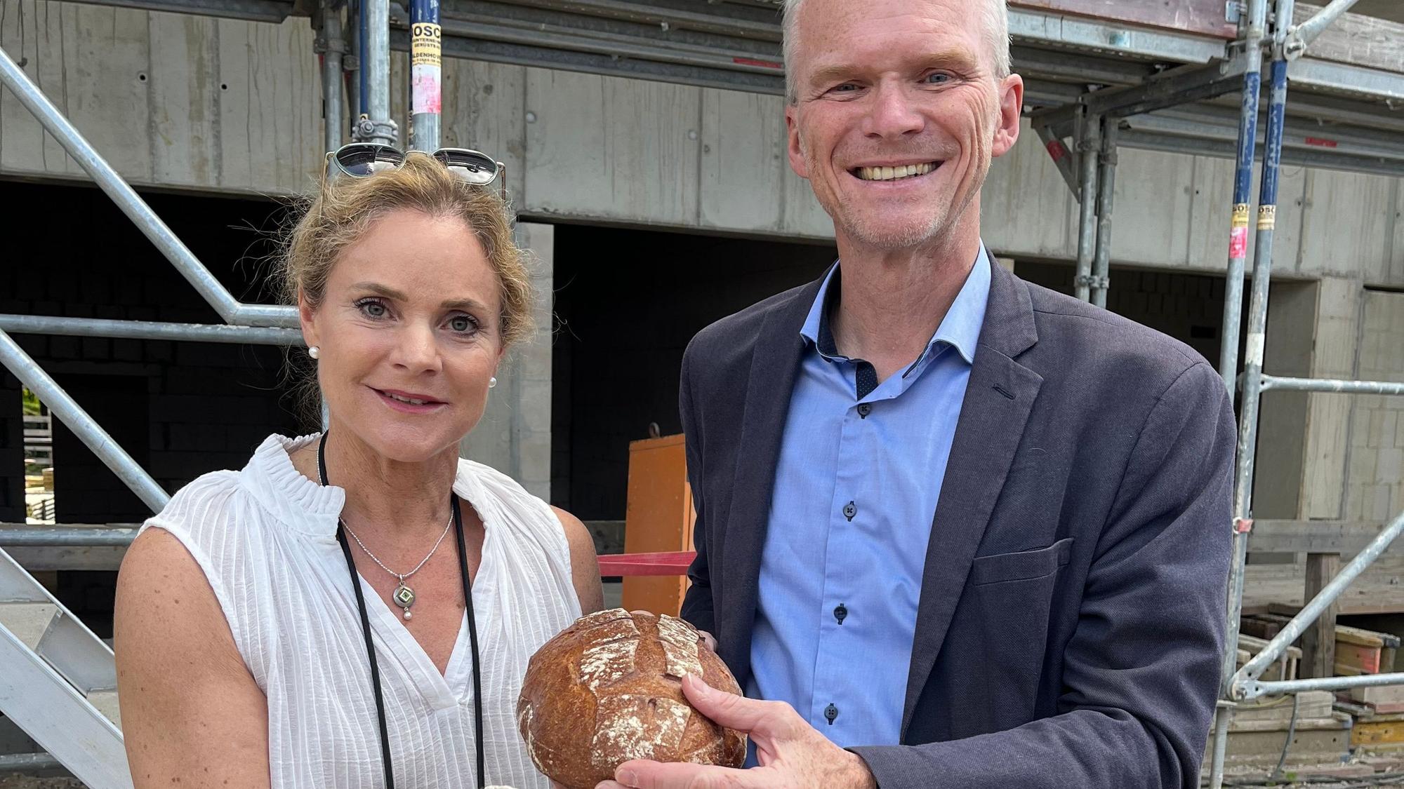 Die beiden stehen nebeneinander. Frau will hält ein Brot in der Hand.