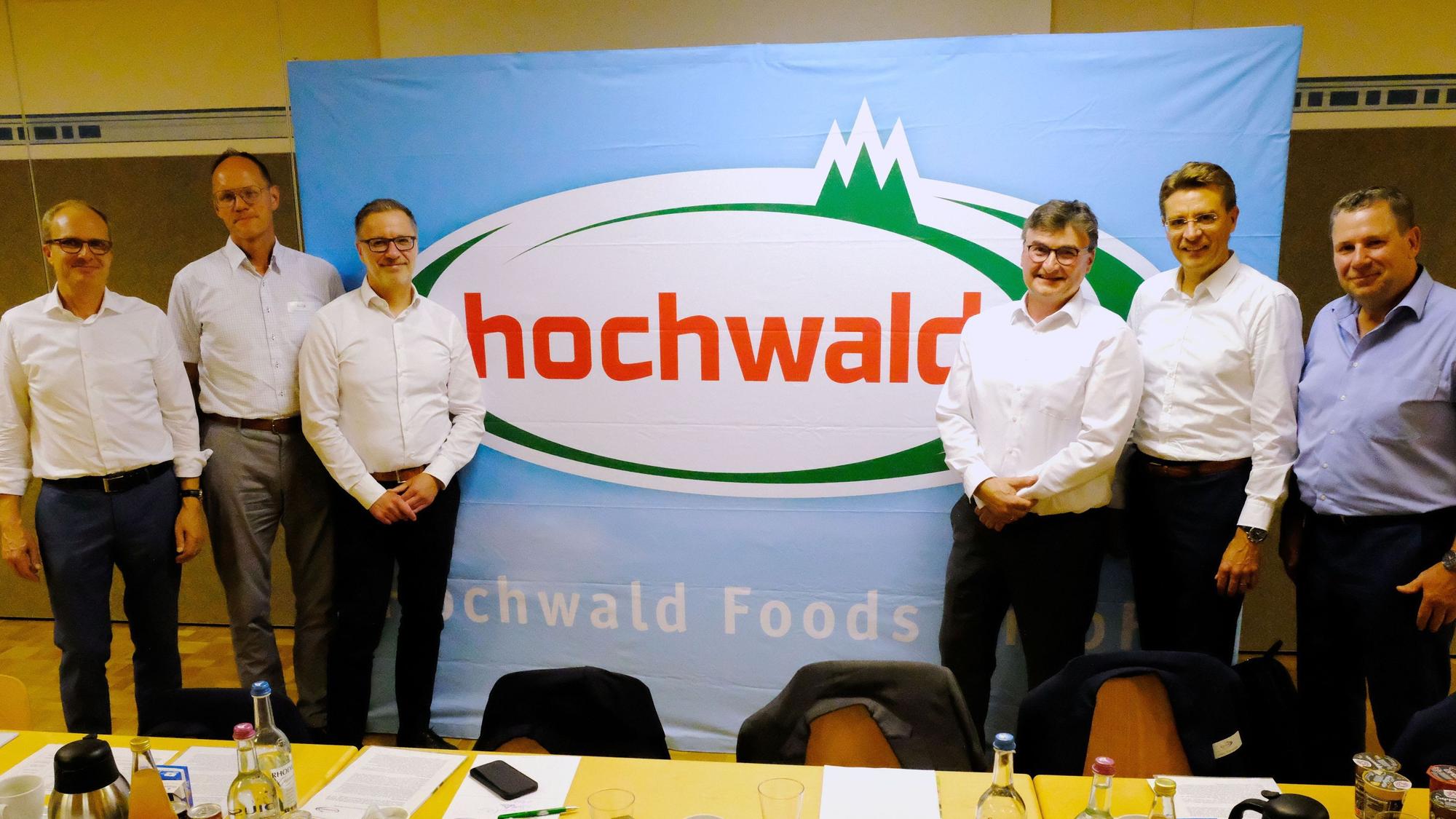 Sechs Männer, fünf im weißen Hemd, einer im blauen, stehen vor einem Plakat der Firma Hochwald.