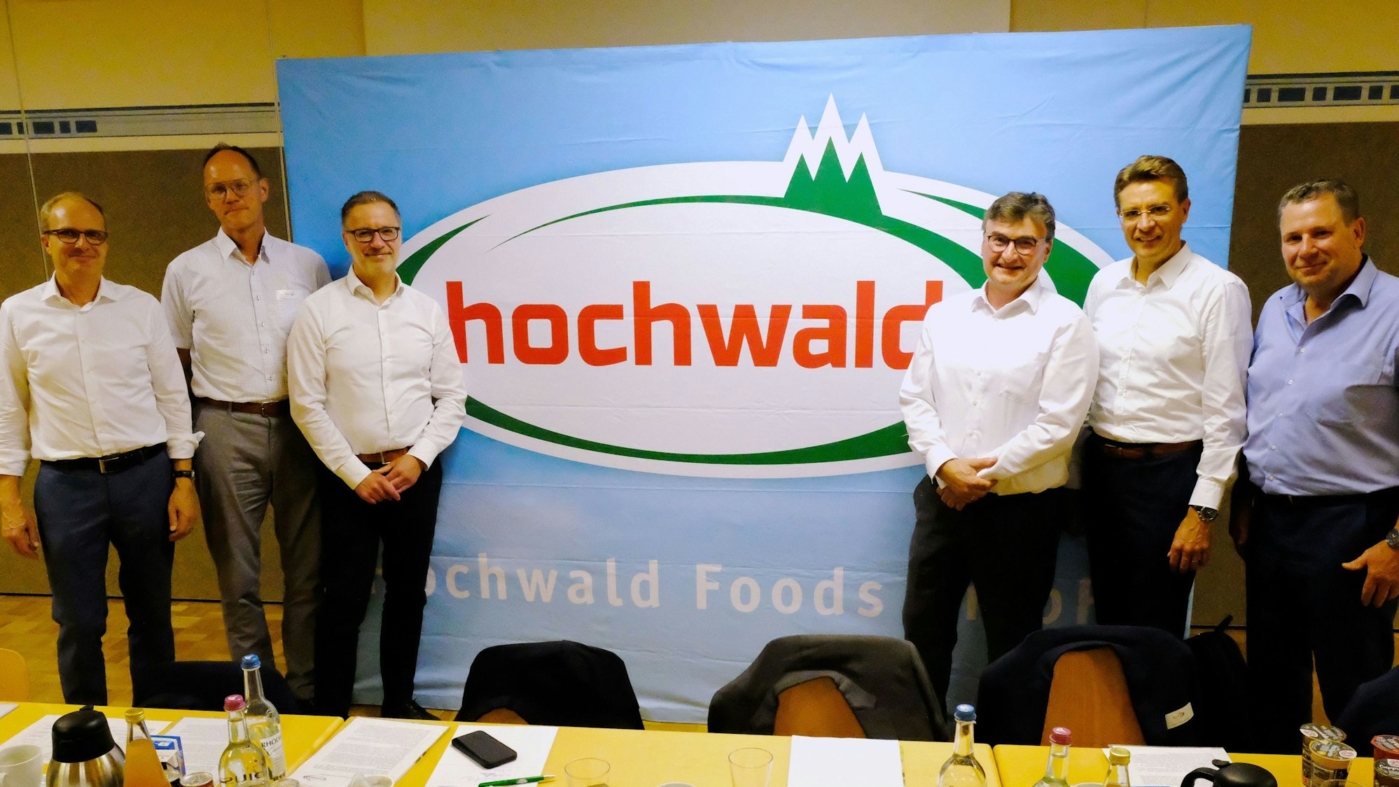 Sechs Männer, fünf im weißen Hemd, einer im blauen, stehen vor einem Plakat der Firma Hochwald.