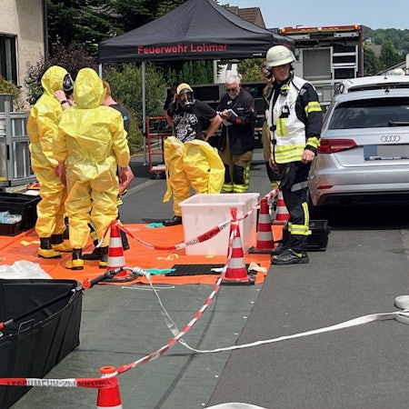 Mehrere Einsatzkräfte in gelben Schutzanzügen stehen auf einer abgesperrten Fläche vor einem Mehrfamilienhaus in Lohmar.