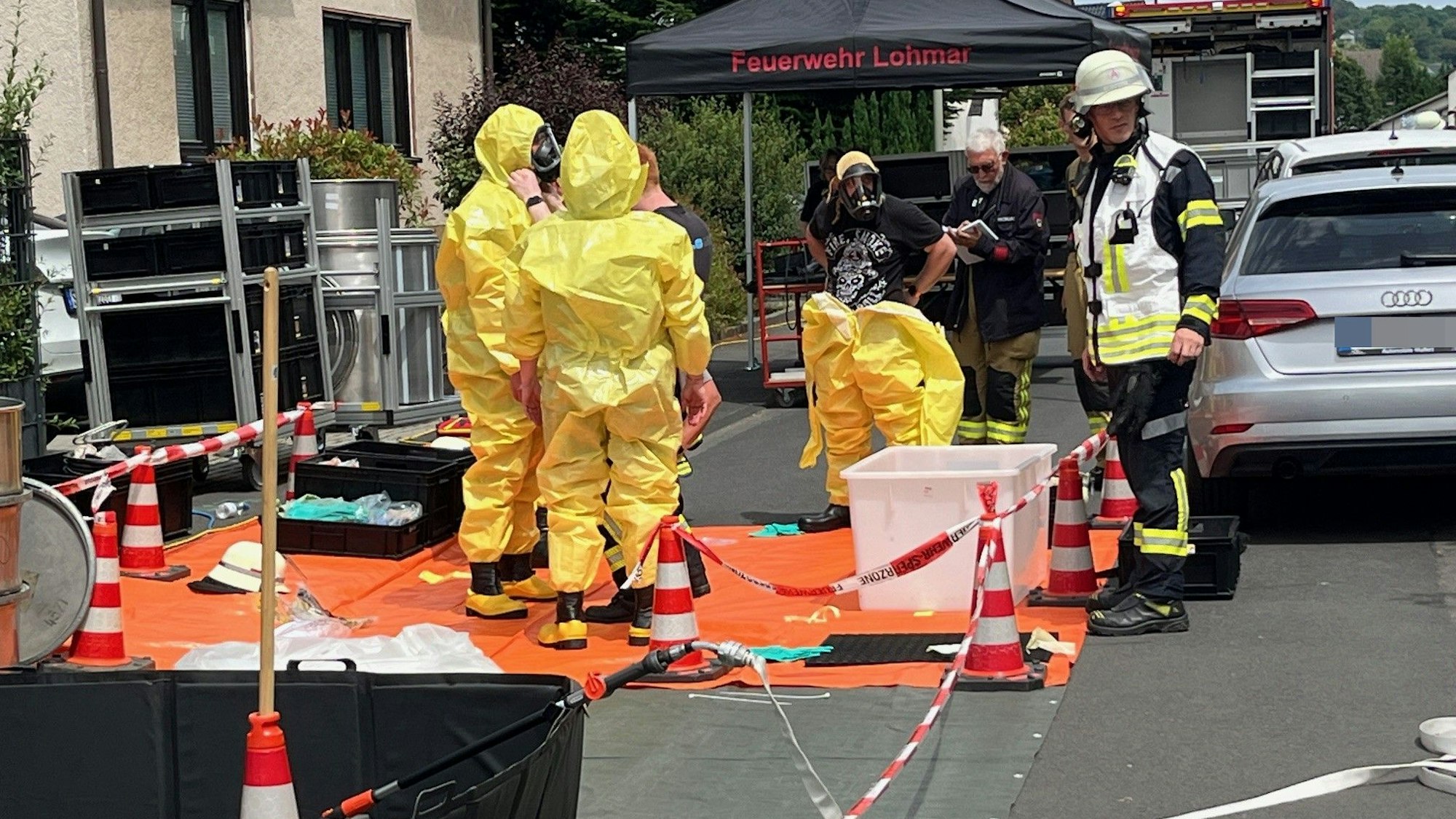 Mehrere Einsatzkräfte in gelben Schutzanzügen stehen auf einer abgesperrten Fläche vor einem Mehrfamilienhaus in Lohmar.