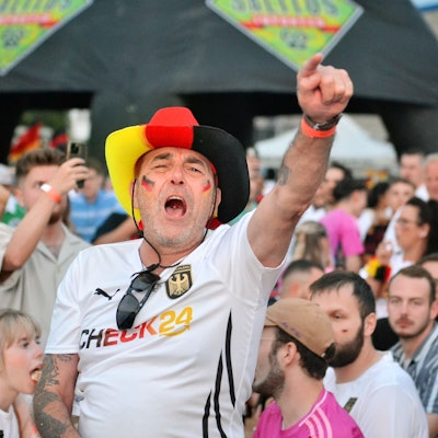 Ein Fan mit Hut in den Deutschlandfarben und mit einem Fan-T-Shirt jubelt und singt beim Public Viewing.