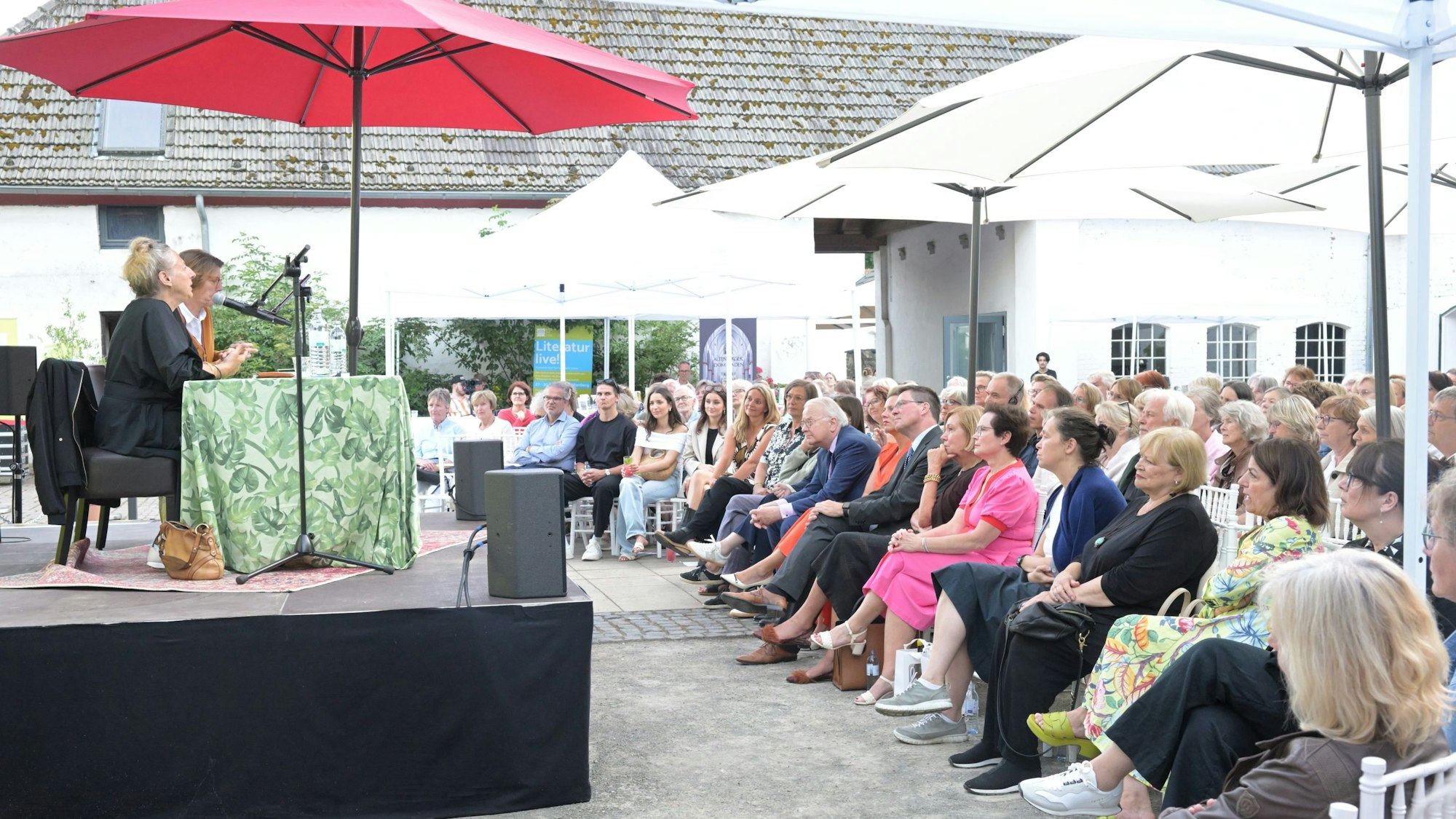Ein Innenhof mit Sonnenschirmen. Auf einer Bühne sitzen zwei Frauen, davor Publikum.