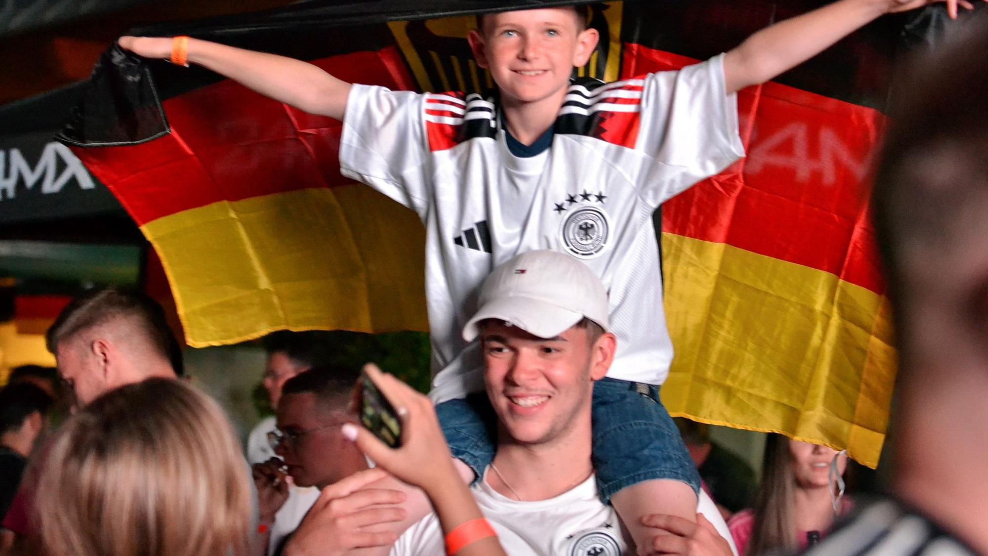 Ein kleiner Junge im Deutschland-Trikot hält eine Deutschland-Fahne hinter sich in die Höhe und sitzt auf den Schultern eines jungen Mannes.