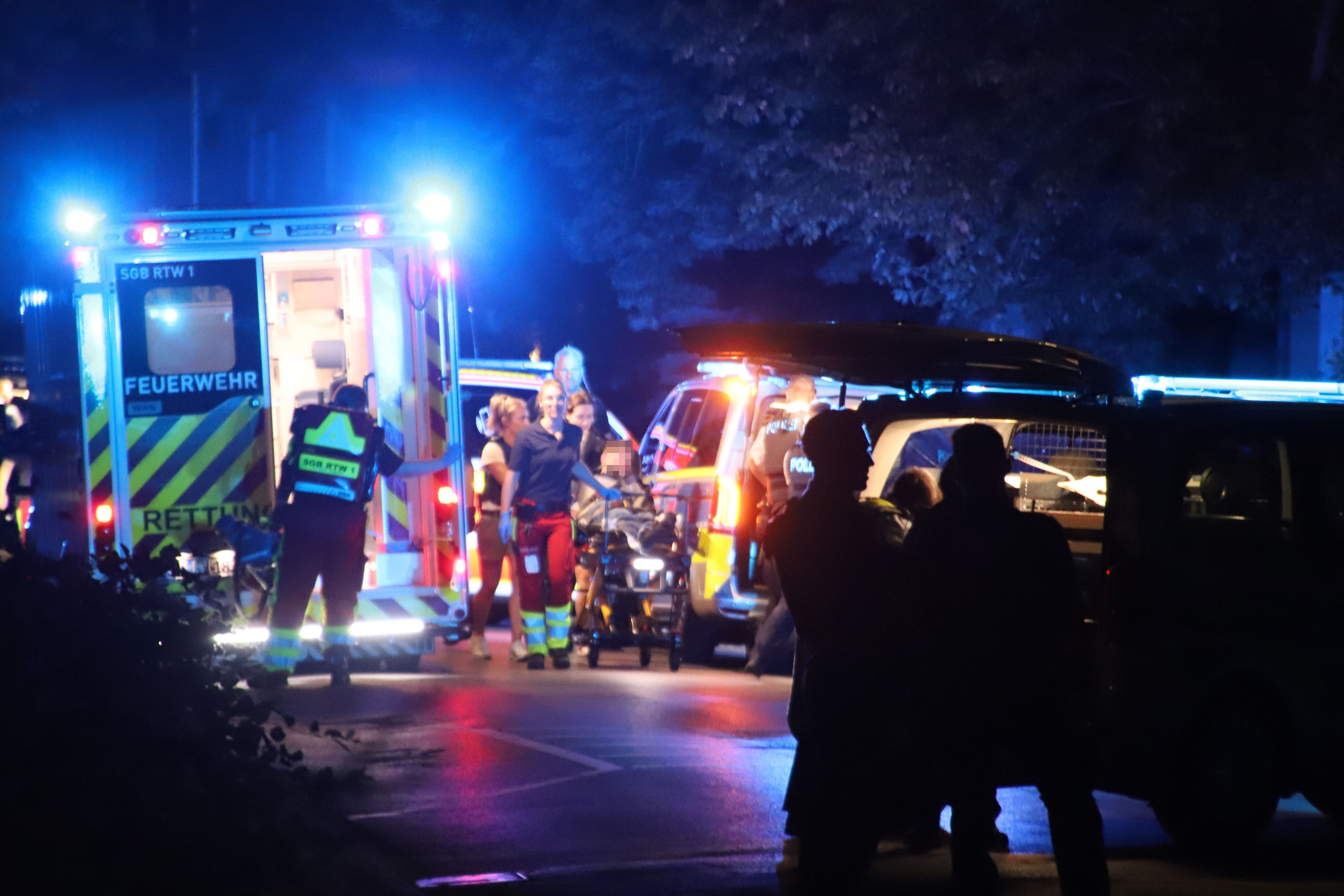 Mehrere Einsatzfahrzeuge von Polizei und Feuerwehr stehen auf einer nächtlichen Straße, Rettungskräfte schieben eine Trage.