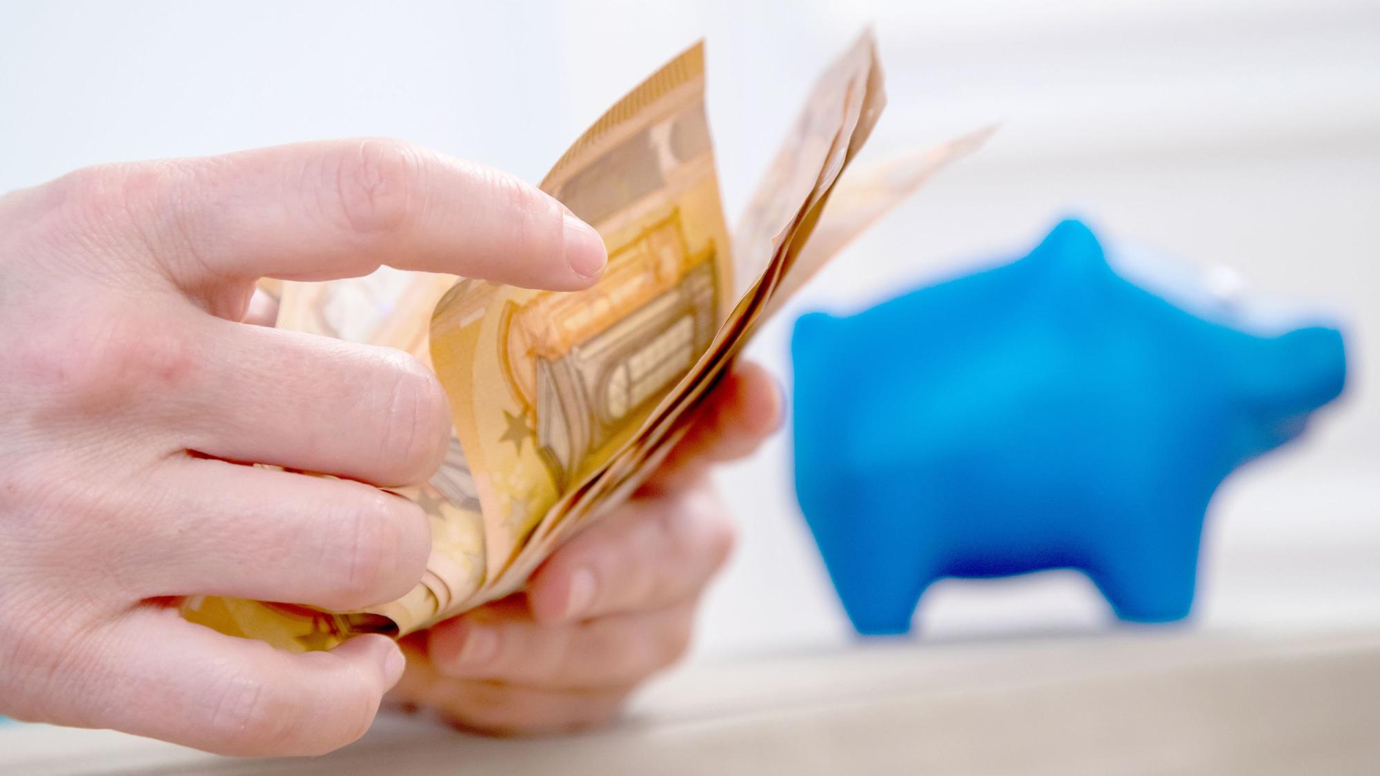 Symbolfoto: Jemand zählt 50-Euro-Scheine ab, im Hintergrund steht ein blaues Sparschwein.