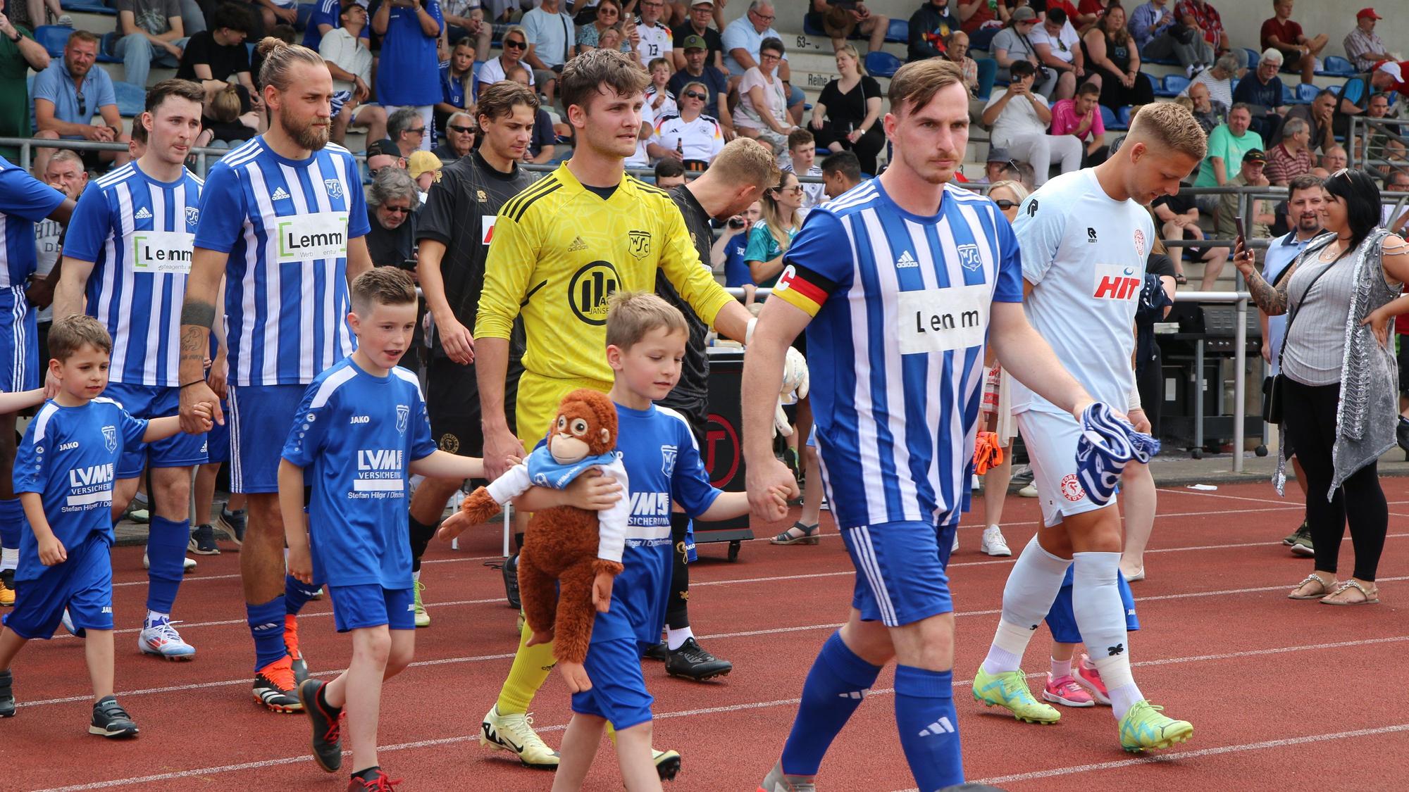 Die Mannschaften gehen aufs Spielfeld. An den Händen halten die Spieler Einlaufkinder. Eines trägt ein Stofftier.