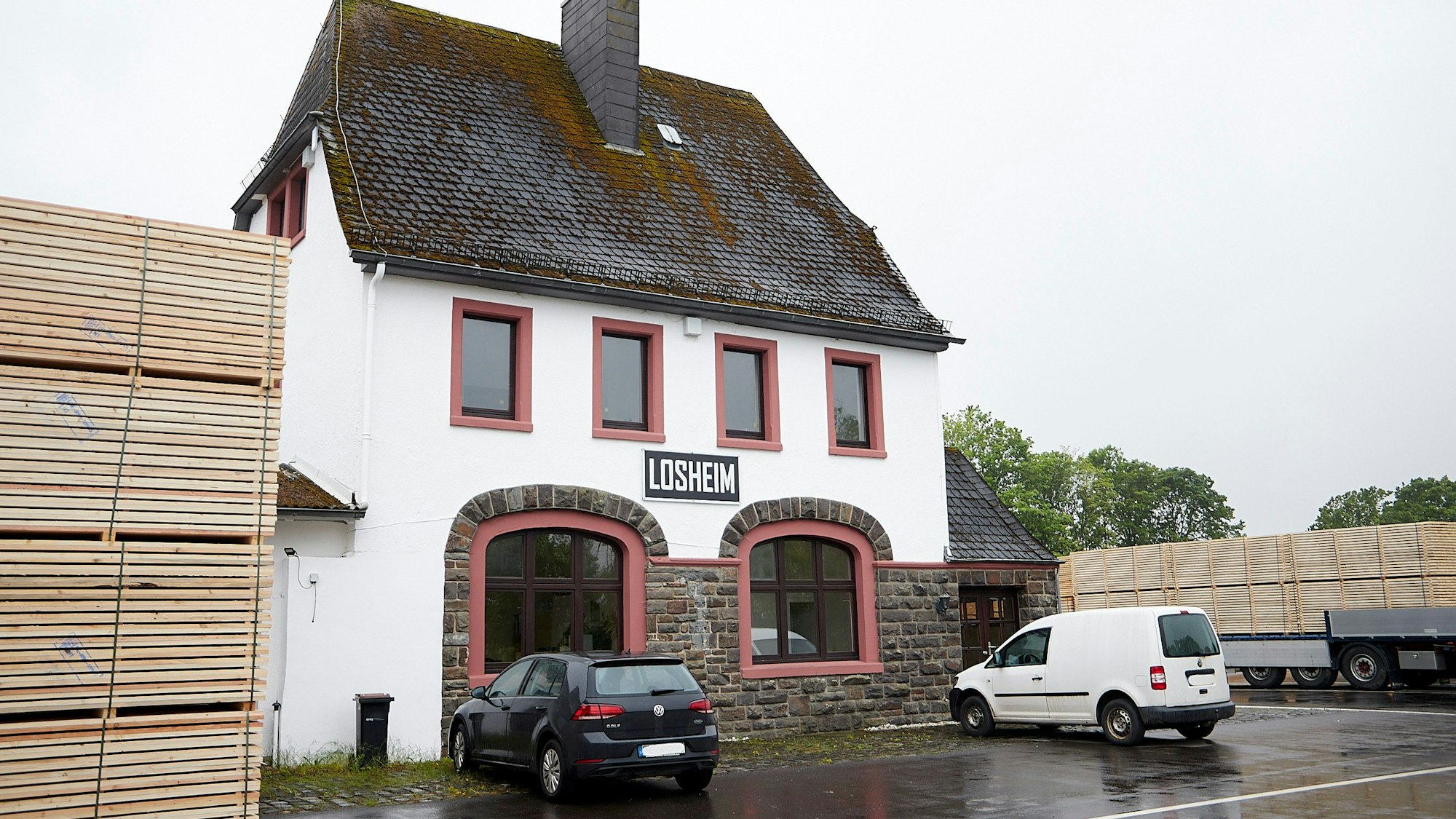Vor dem alten Losheimer Bahnhof parken zwei Autos. Links ist ein Stapel mit Schnittholz zu sehen.