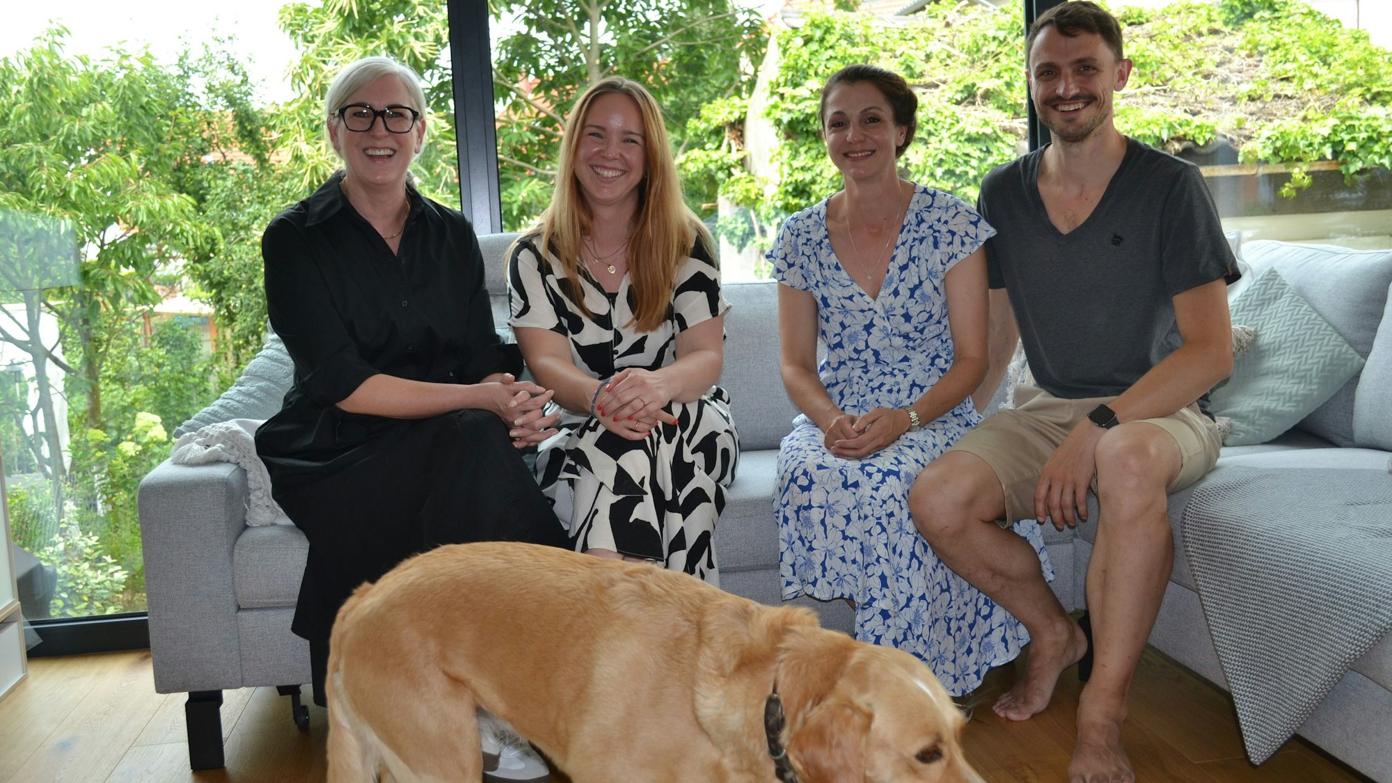Drei Frauen und ein Mann sitzen auf einer Couch, vor ihnen steht ein Hund.