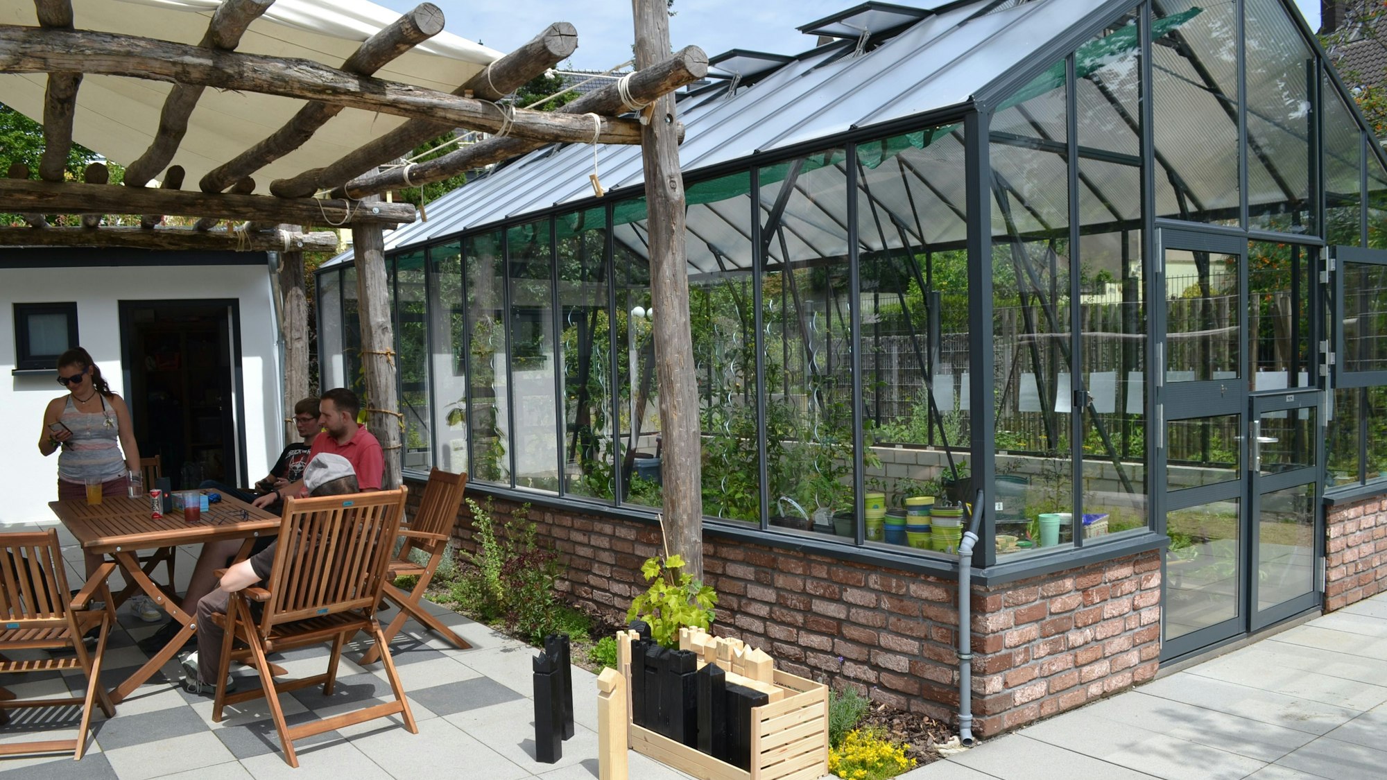Tag der Architektur im Rhein-Sieg-Kreis: Menschen sitzen auf Gartenmöbeln unter einem Sonnendach aus Baumstämmen, daneben steht ein großes Gewächshaus.