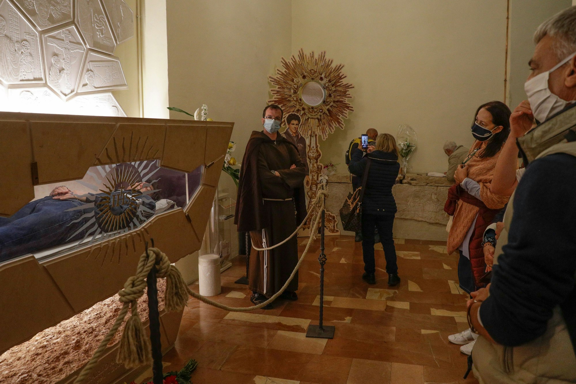 Merhere Personen mit Gesichtsmaksen stehen neben einem einsehbaren Glassarg, in dem ein junger Mannliegt. (Archivbild)