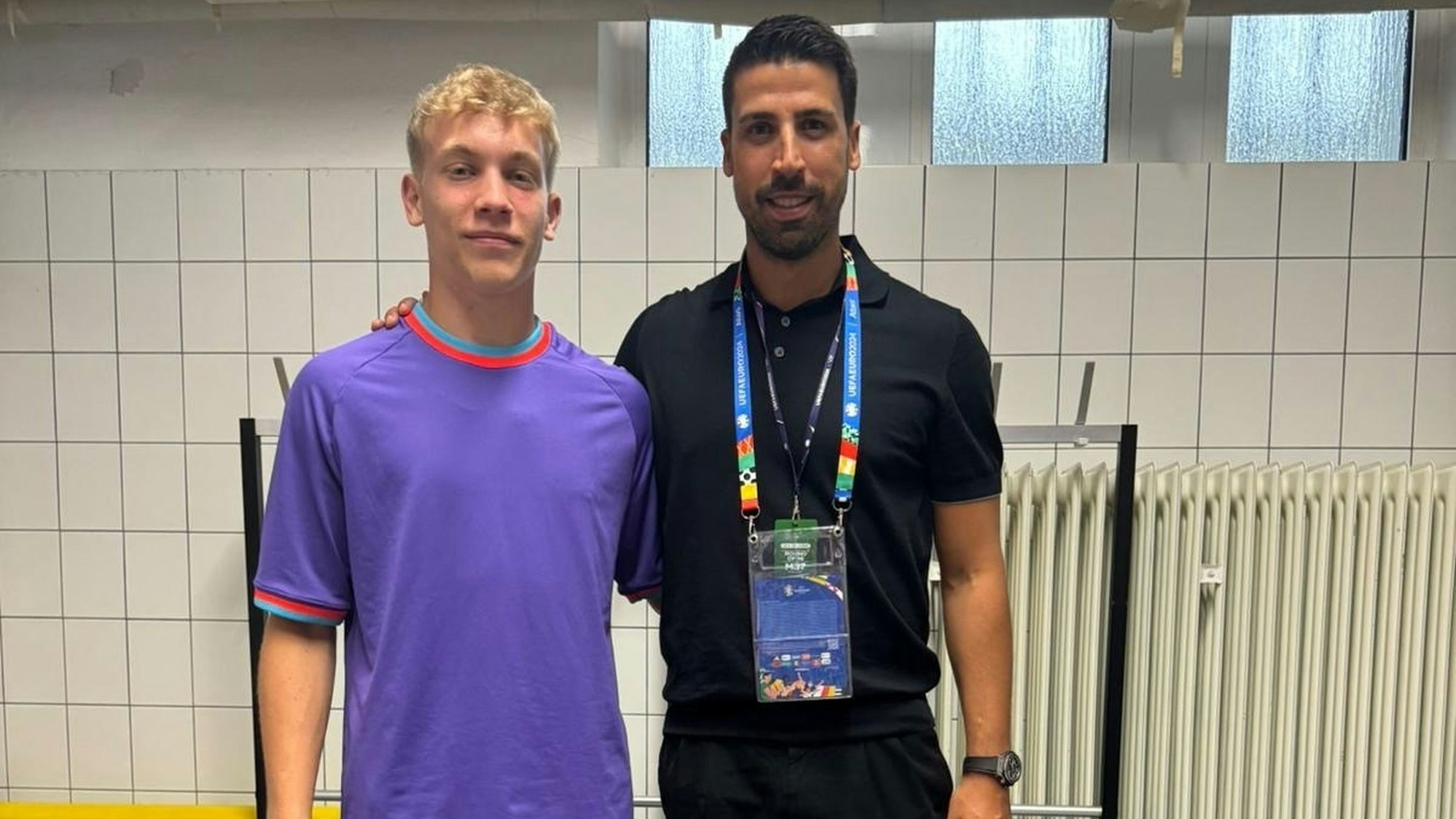 Der Kispenicher Julian Seliger trägt ein lilafarbenes Shirt. Er steht in einer Umkleidekabine Arm in Arm mit Weltmeister Sami Khedira.