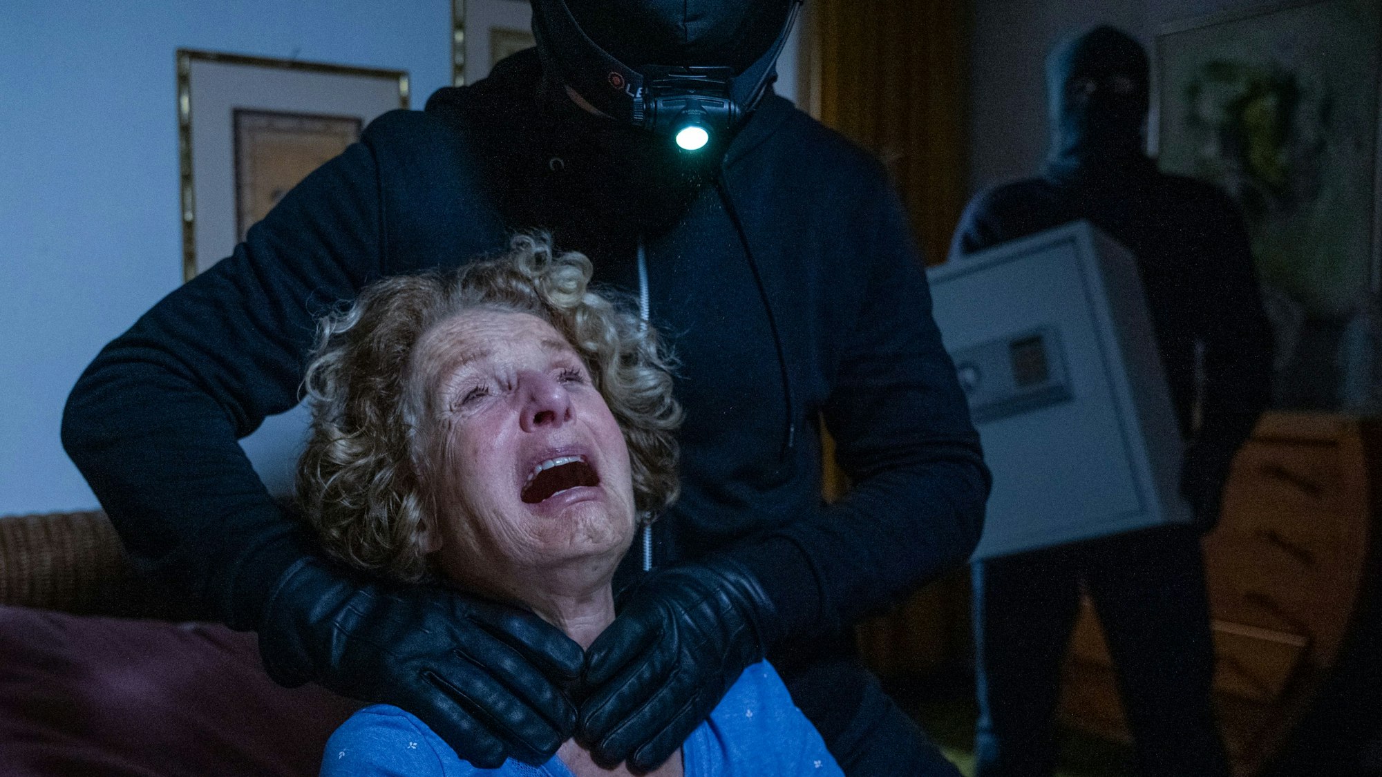 Eine Szene aus dem Film: Das Opfer wird von einem maskierten Mann am Hals gepackt, im Hintergrund steht ein ebenfalls maskierter Mann mit einem Tresor.