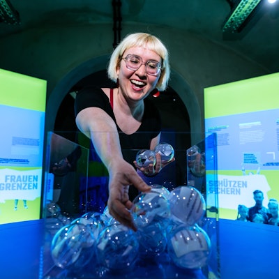 Kuratorin Rebekka Blum greift in eine Vase mit durchsichtigen Kugeln in denen Zettel sind.