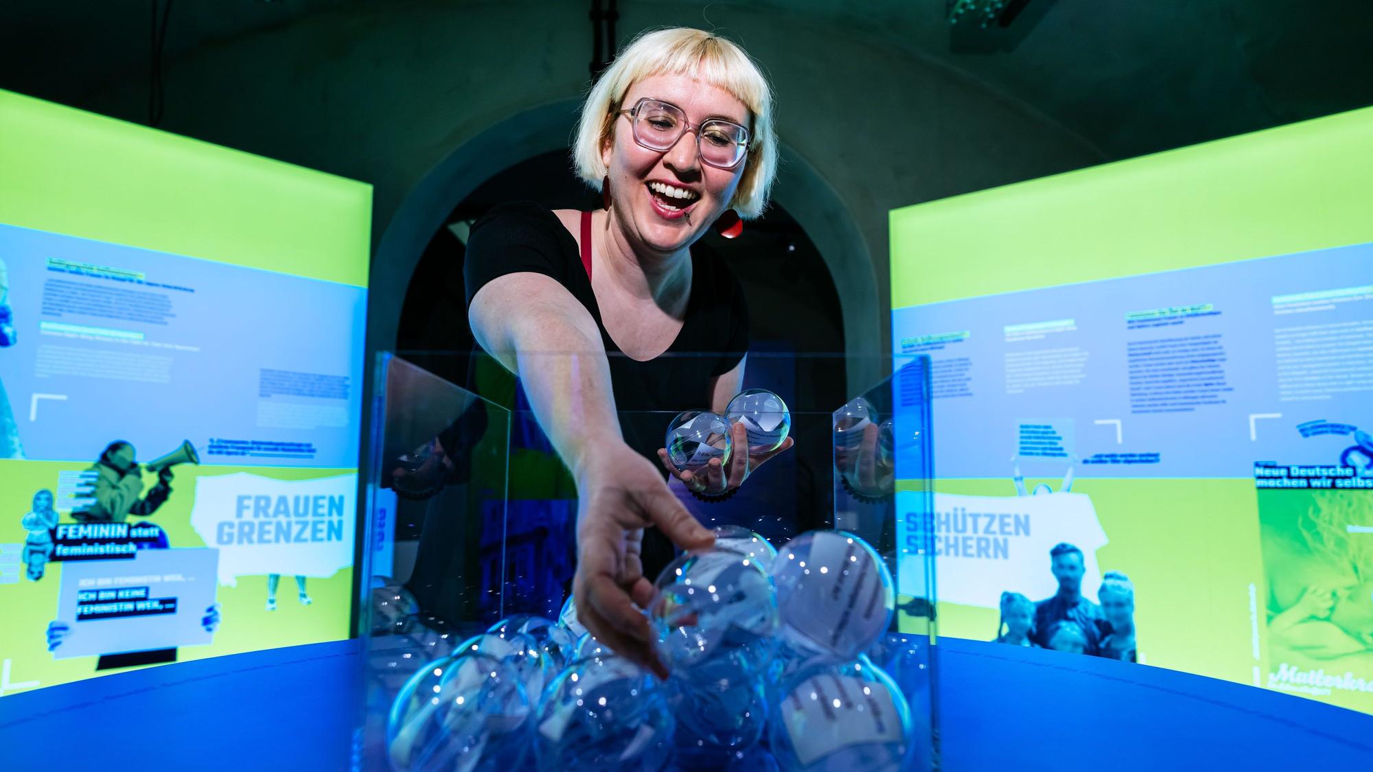 Kuratorin Rebekka Blum greift in eine Vase mit durchsichtigen Kugeln in denen Zettel sind.