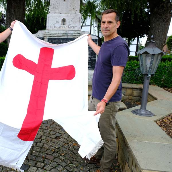Bernd Abel (l.) und Christof Jansen mit der zerstörten Fahne vor dem Kriegerdenkmal in der Ortsmitte von Wachendorf. Die Fahne ist weiß und hat in der Mitte ein großes rotes Kreuz.