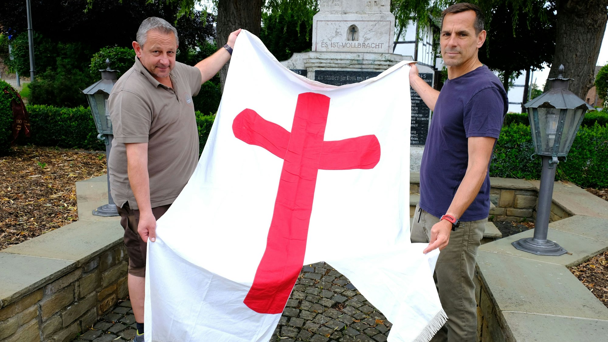 Bernd Abel (l.) und Christof Jansen mit der zerstörten Fahne vor dem Kriegerdenkmal in der Ortsmitte von Wachendorf. Die Fahne ist weiß und hat in der Mitte ein großes rotes Kreuz.
