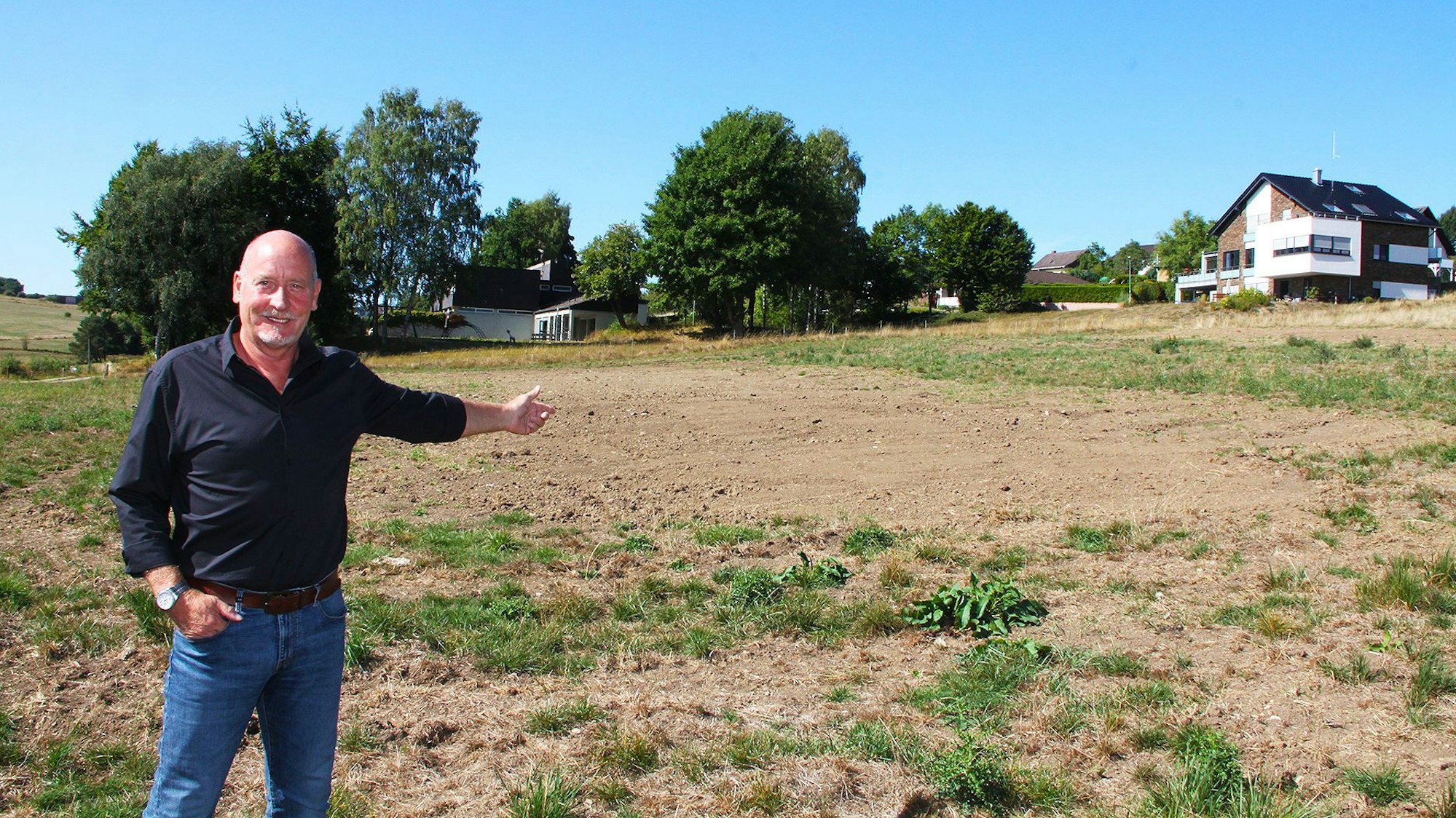 Ein Mann in Jeans und schwarzem Hemd zeigt auf eine Wiese, auf der künftig Häuser gebaut werden sollen. Im Hintergrund sind bereits Häuser zu sehen.