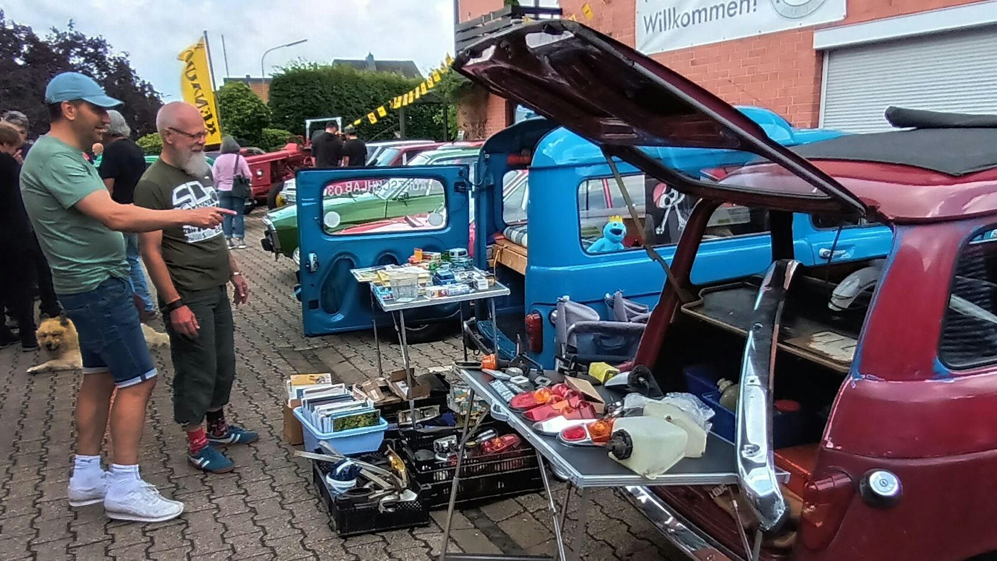 In und vor einem offenen Kofferraum präsentiert ein Mann Ersatzteile.