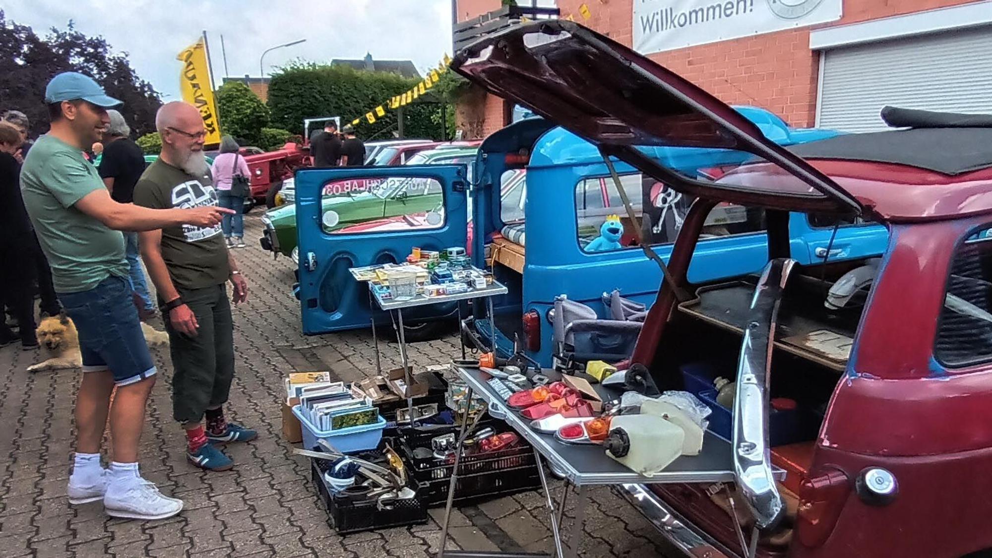In und vor einem offenen Kofferraum präsentiert ein Mann Ersatzteile.