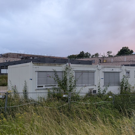Das Foto zeigt Baucontainer auf einem verwilderten Grundstück vor der Baustelle der Feuerwache in Hermülheim.