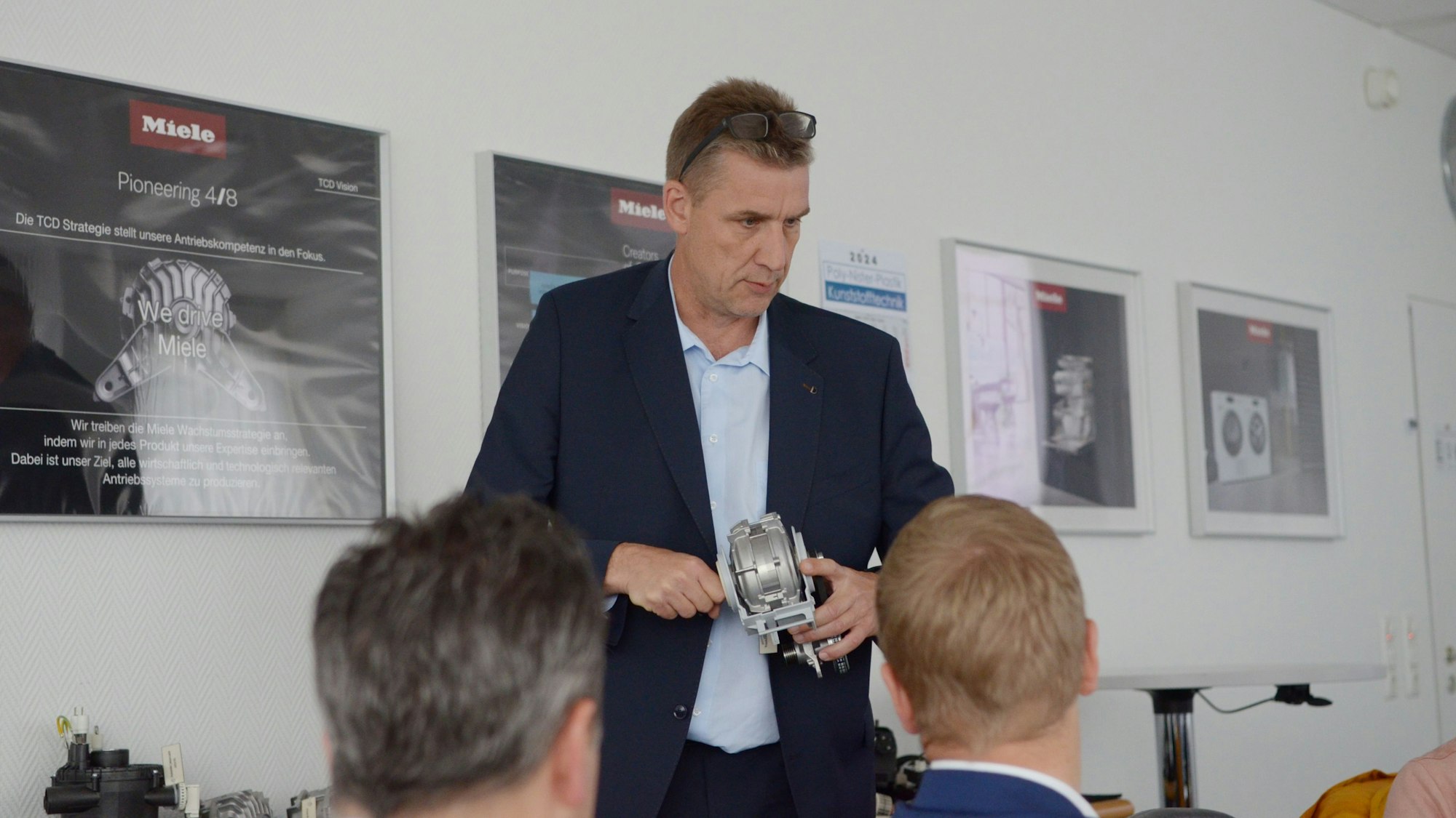 Hendrik Krämer hat einen Motor in der Hand, hinter ihm an der Wand Plakate mit Miele-Produkten.