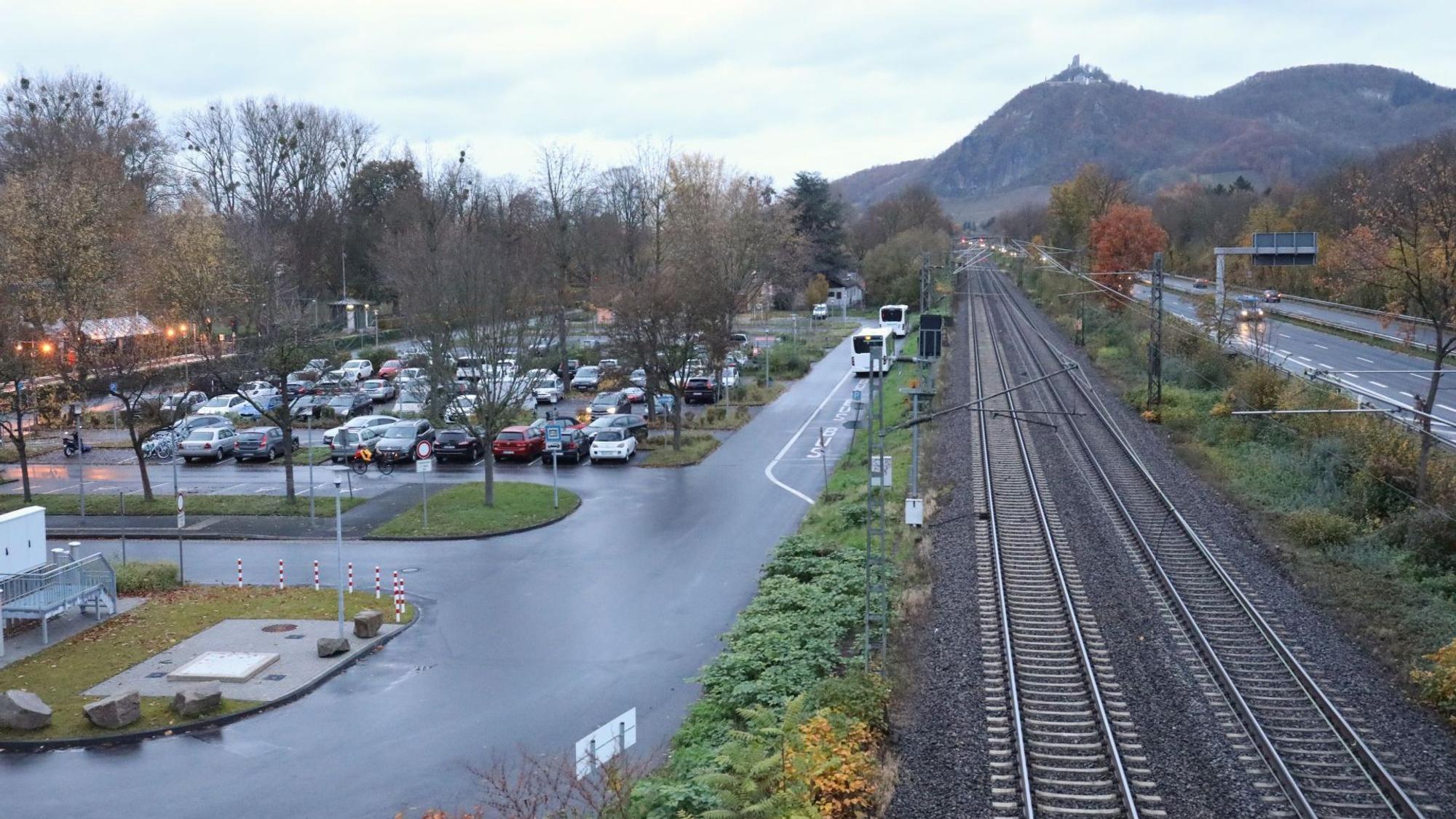Blick auf einen Parkplatz und Bahngleise sowie eine Autobahn. Im Hintergrund Berge und eine Burg.