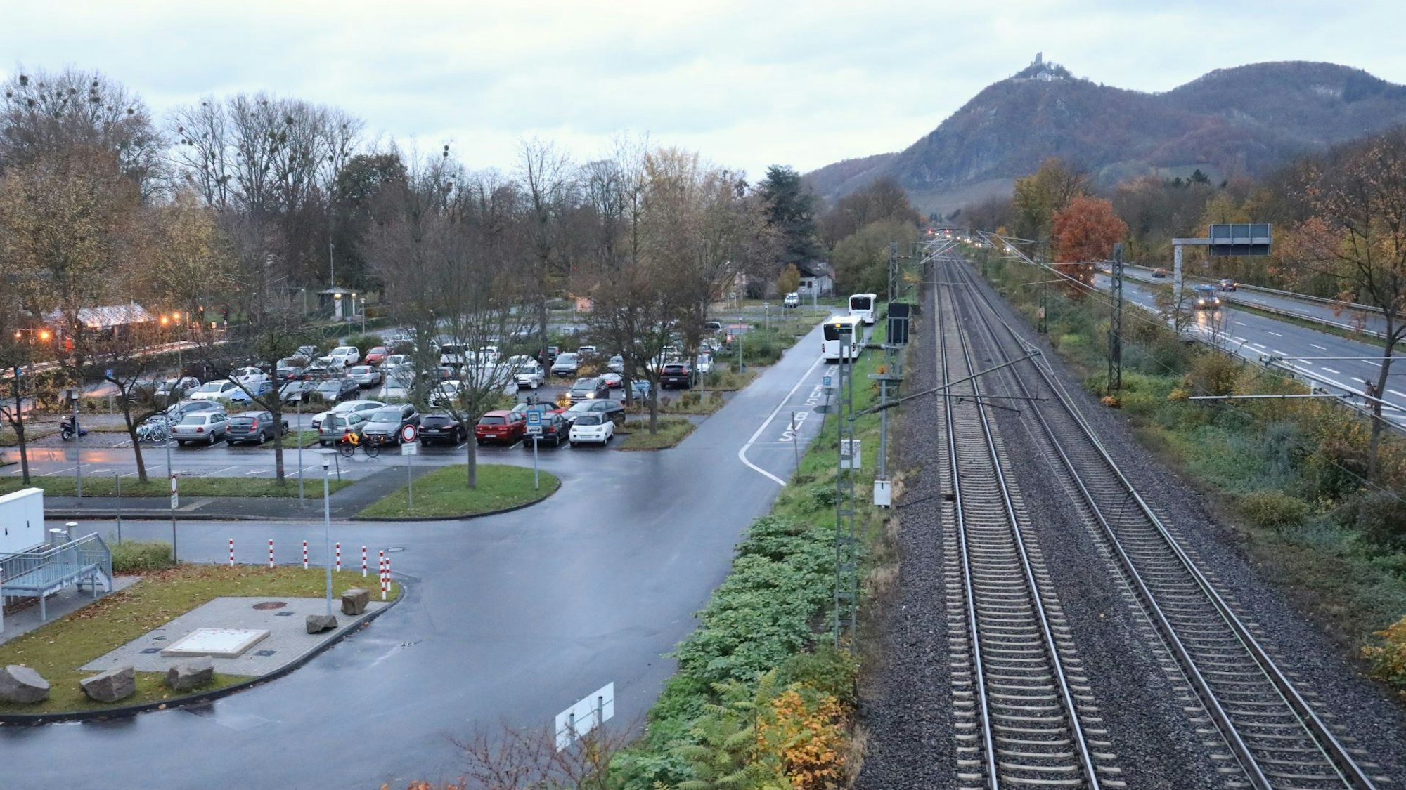Blick auf einen Parkplatz und Bahngleise sowie eine Autobahn. Im Hintergrund Berge und eine Burg.