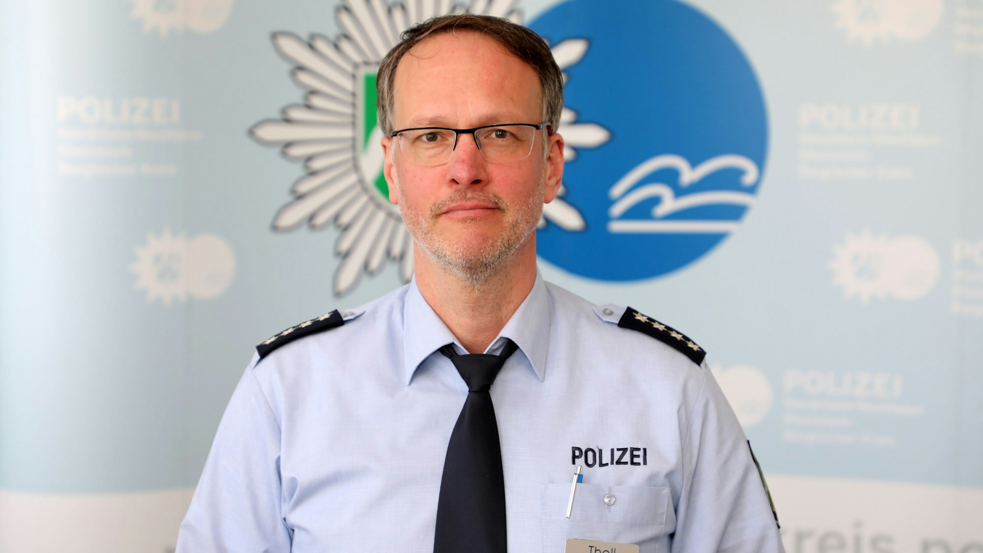Rhein-Bergs Polizeisprecher Christian Tholl steht vor einem Logo seiner Polizeibehörde.
