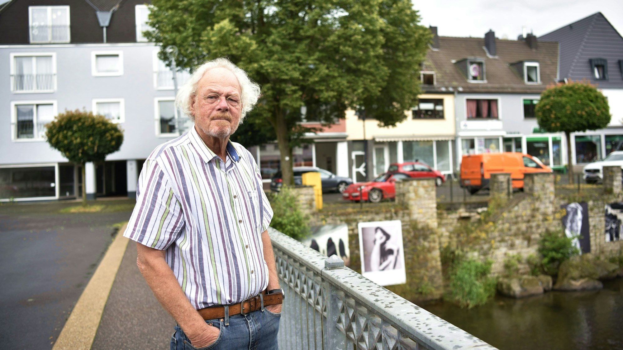 Ein Mann mit längeren weißen Haaren und kurzärmeligen, gestreiften Hemd steht auf einer Brücke, im Hintergrund sind Häuser und Bäume zu erkennen.