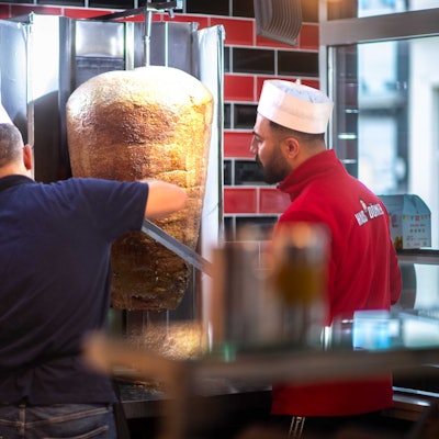 Das Bild zeigt zwei Mitarbeiter, die von zwei Döner-Spießen Fleisch abschneiden.