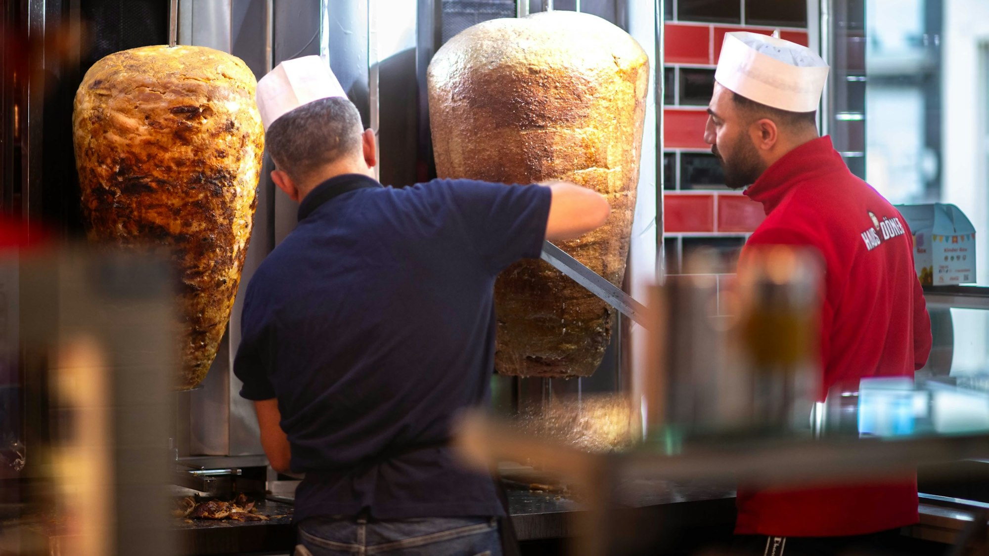 Das Bild zeigt zwei Mitarbeiter, die von zwei Döner-Spießen Fleisch abschneiden.