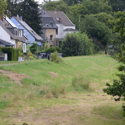 Hinter einem mit Gras bewachsenen Flussdeich stehen Wohnhäuser.