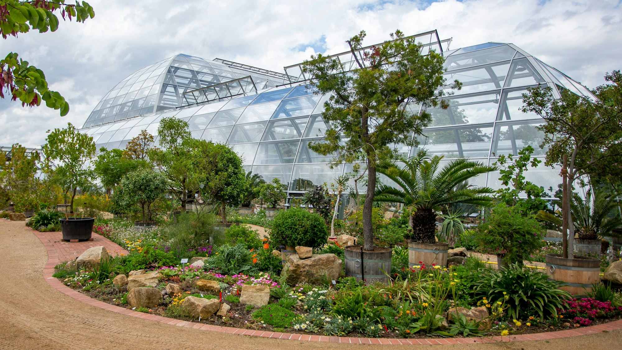 Auch von außen schön: Die Schaugewächshäuser mit umgebender Bepflanzung. Blumenrabatten und halbhohe Büsche zwischen Natursteinen schmücken die Umgebung