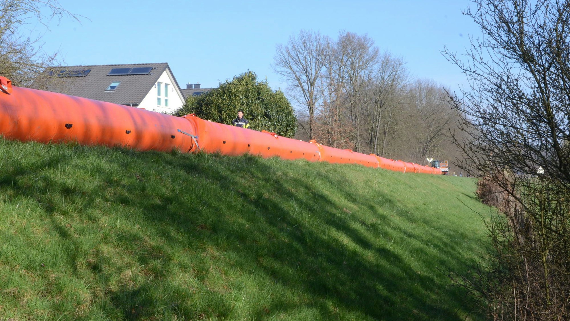 Ein mit Gras bewachsener Flussdeich, auf der Deichkrone liegen große orange Schläuche. Hinter dem Deich ragt ein Dachfirst heraus.