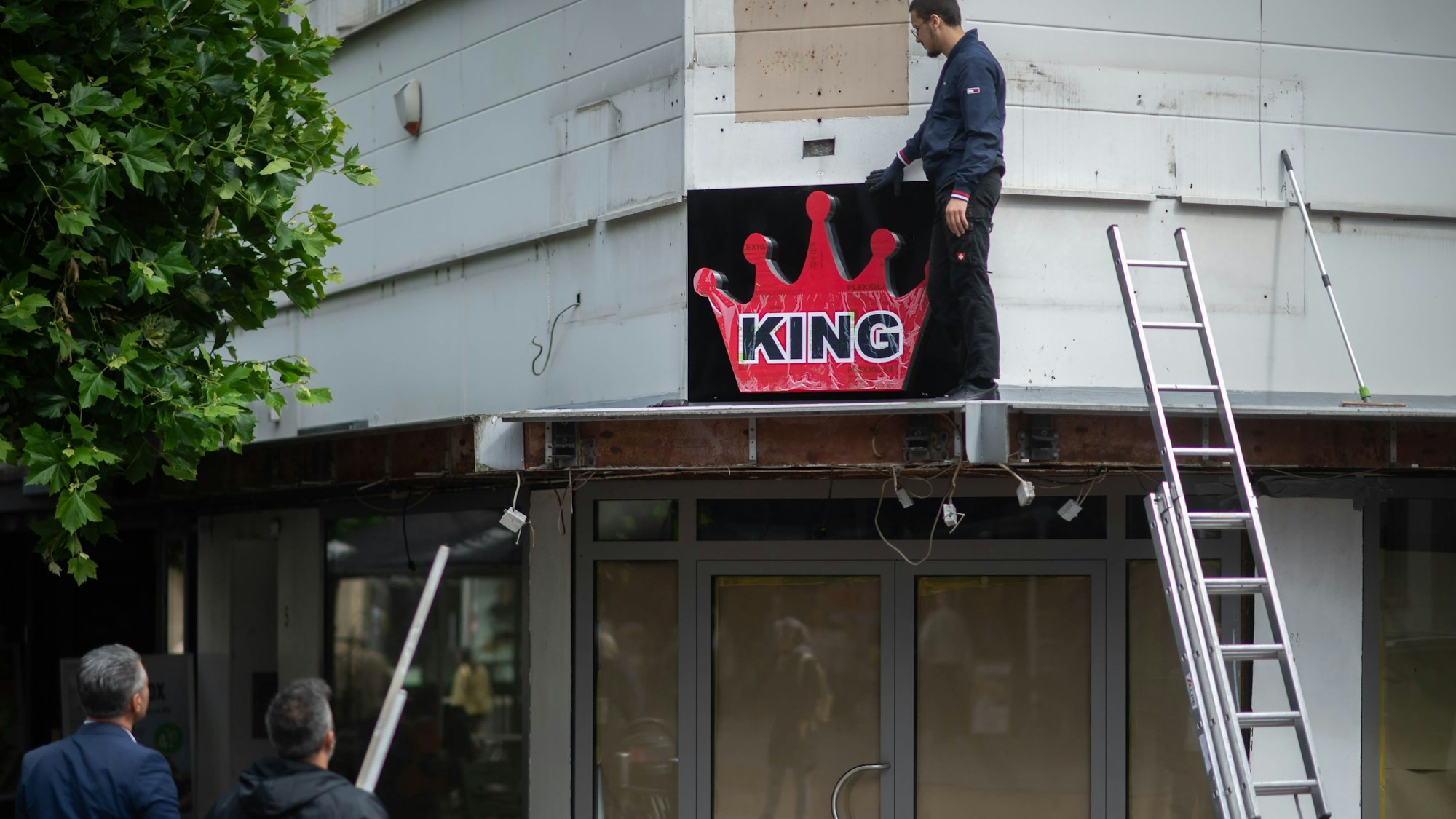 Das Bild zeigt einen Mann, der auf dem Vordach eines Geschäfts steht. Er platziert ein Reklame-Schild mit der Aufschrift „King“.