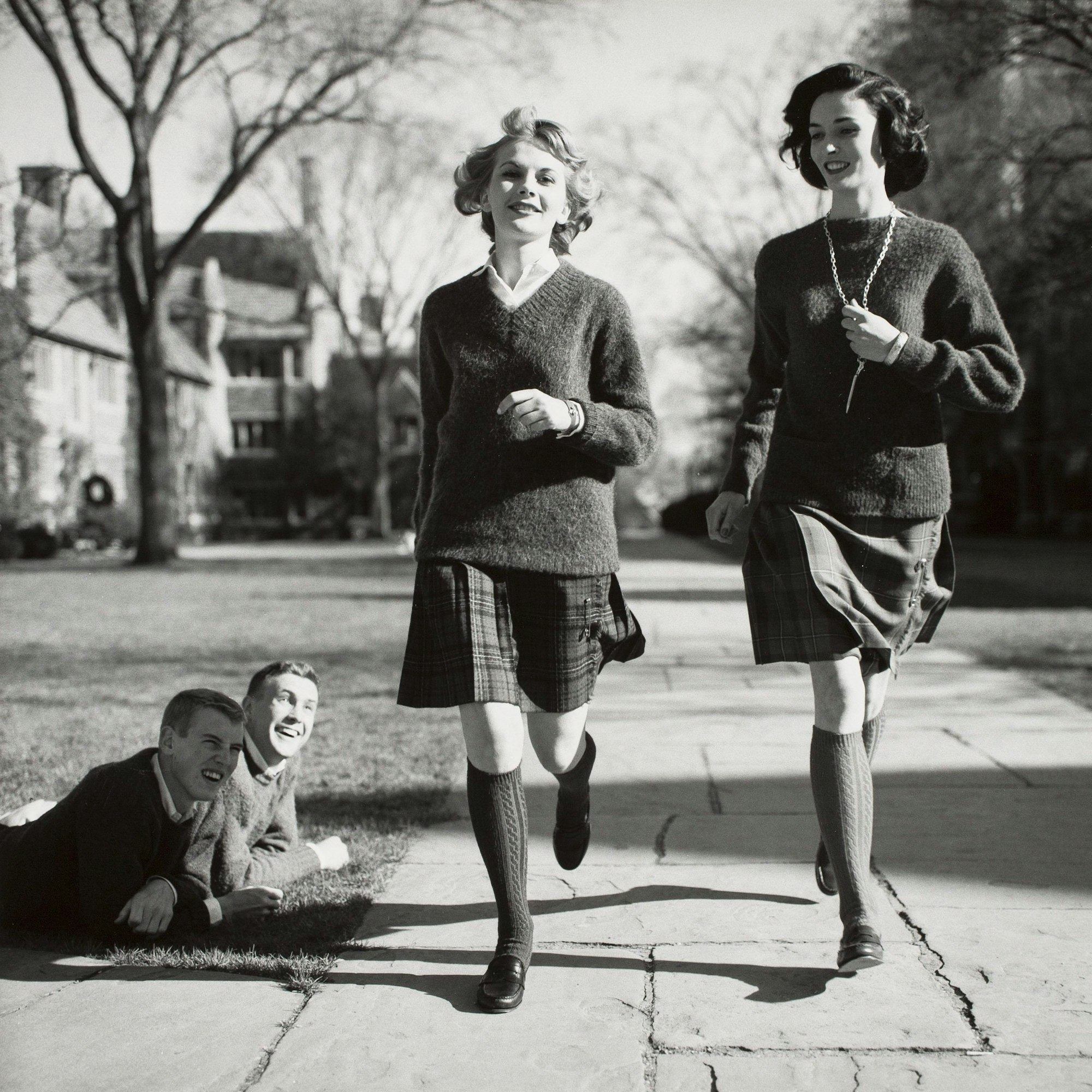 Die Schwarz-weiß-Aufnahme von 1958 zeigt zwei junge Frauen, die Kniestrümpfe, Röcke und Pullover tragen und an zwei Männern vorbeigehen, die auf einer Rasenfläche liegen und ihnen hinterherschauen.
