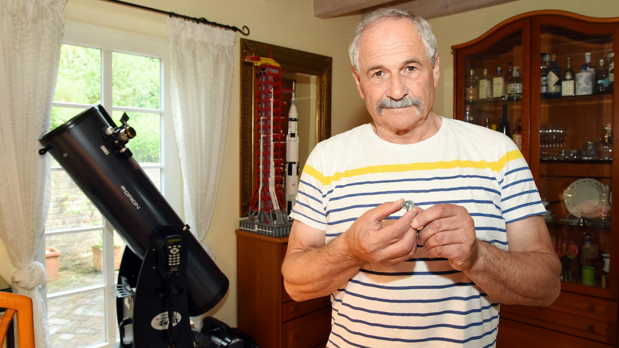 Michael Stumpe hält ein winziges Mondstück zwischen seinen Fingern. Im Hintergrund ist ein Teleskop zu sehen.