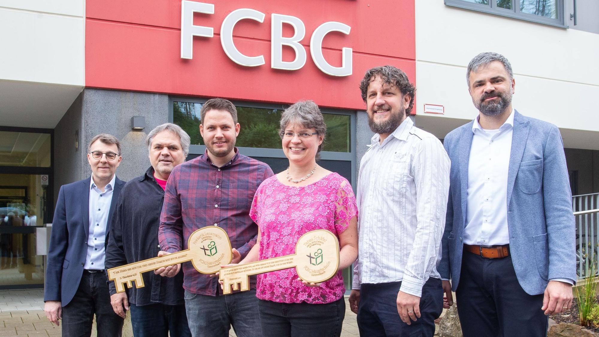 Schlüsselübergabe bei der FCBG-Hauptschule (v.l.): Jakob Holzrichter (Schulträger), Ex-Schulleiter Thomas Sieling, Jan-Frederik Lessmann (neuer Schulleiter), Tabea Schäfer (neue stellvertretende Schulleiterin), Ralf Sassenhausen (ehemaliger stellvertretender Schulleiter), Viktor Pritzkau (Schulträger) stehen nebeneinander. Sie halten zwei große Schlüssel in den Händen.